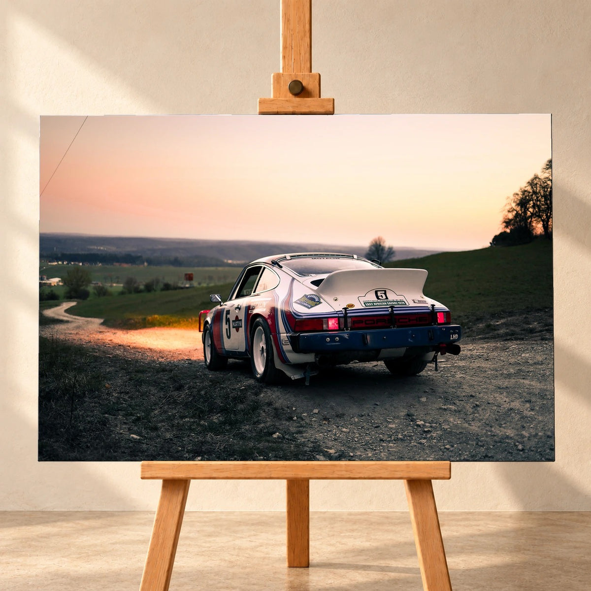 Frontale Visualisierung eines Leinwandbilds im Querformat auf einer Holzstaffelei vor heller Wand. Das Motiv zeigt einen weißen Porsche 911 Safari mit rot-blauen Streifen und Startnummer 5 schräg von hinten auf einem Schotterweg unter orange-rosa Abendhimmel.