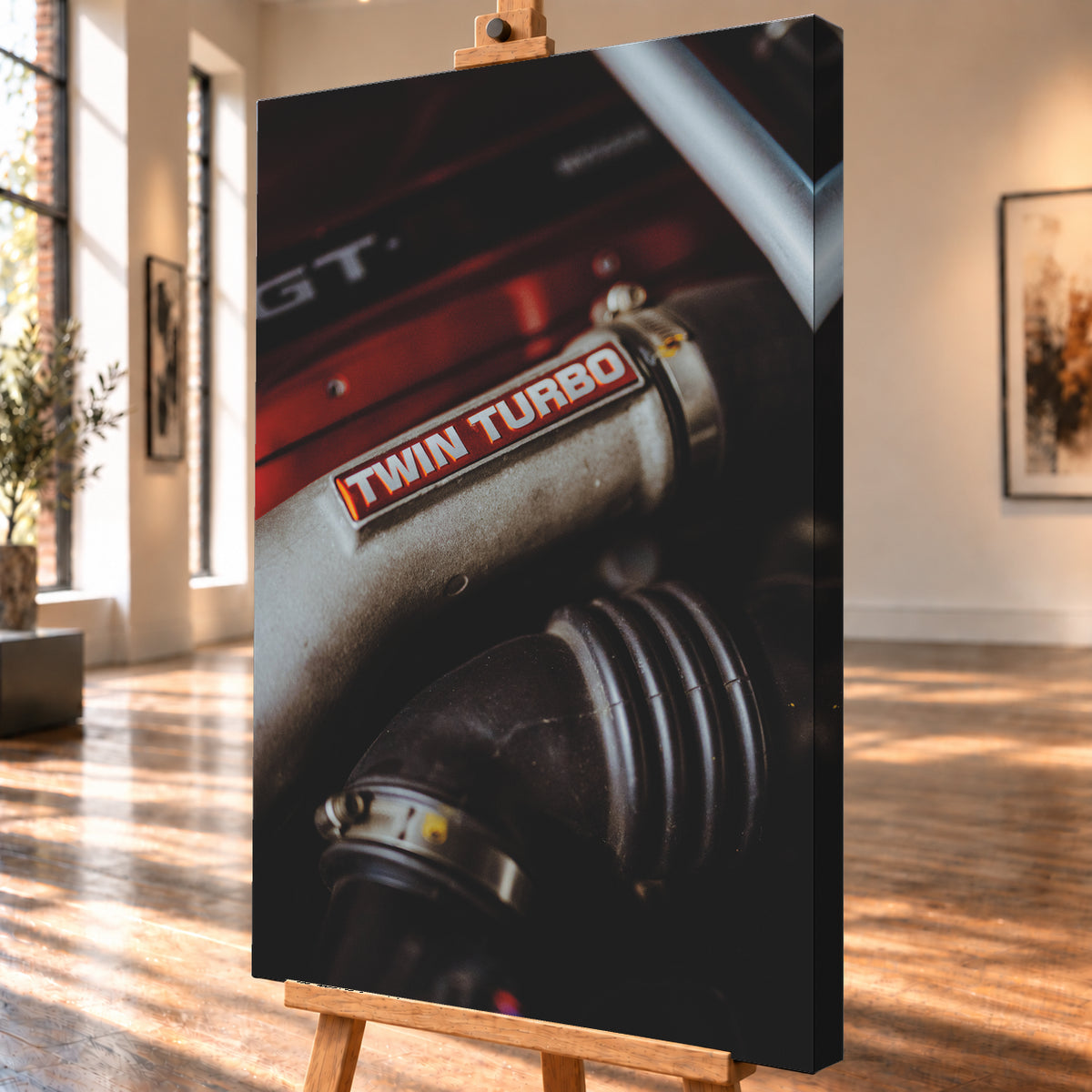 Detailaufnahme eines Motorraums mit metallisch grauem Bauteil und rot-weißem „TWIN TURBO“-Schriftzug. Schwarze gerippte Schläuche, rote Flächen, ein helles diagonales Element und geringe Schärfentiefe prägen das dunkle Bild; Visualisierung als Leinwand-Wandbild, schräg von der Seite auf einer Holzstaffelei.