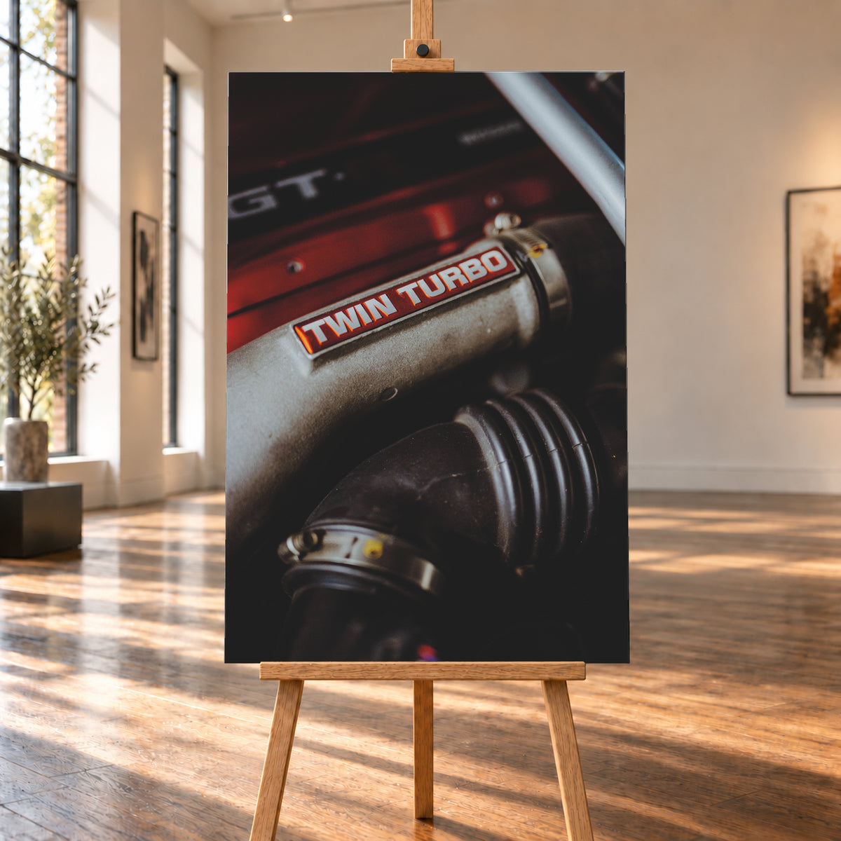 Detailaufnahme eines Motorraums mit metallisch grauem Bauteil und rot-weißem „TWIN TURBO“-Schriftzug. Schwarze gerippte Schläuche, rote Flächen, ein helles diagonales Element und geringe Schärfentiefe prägen das dunkle Bild; Visualisierung als Leinwand-Wandbild, auf einer Holzstaffelei in einem hellen Galerieraum.