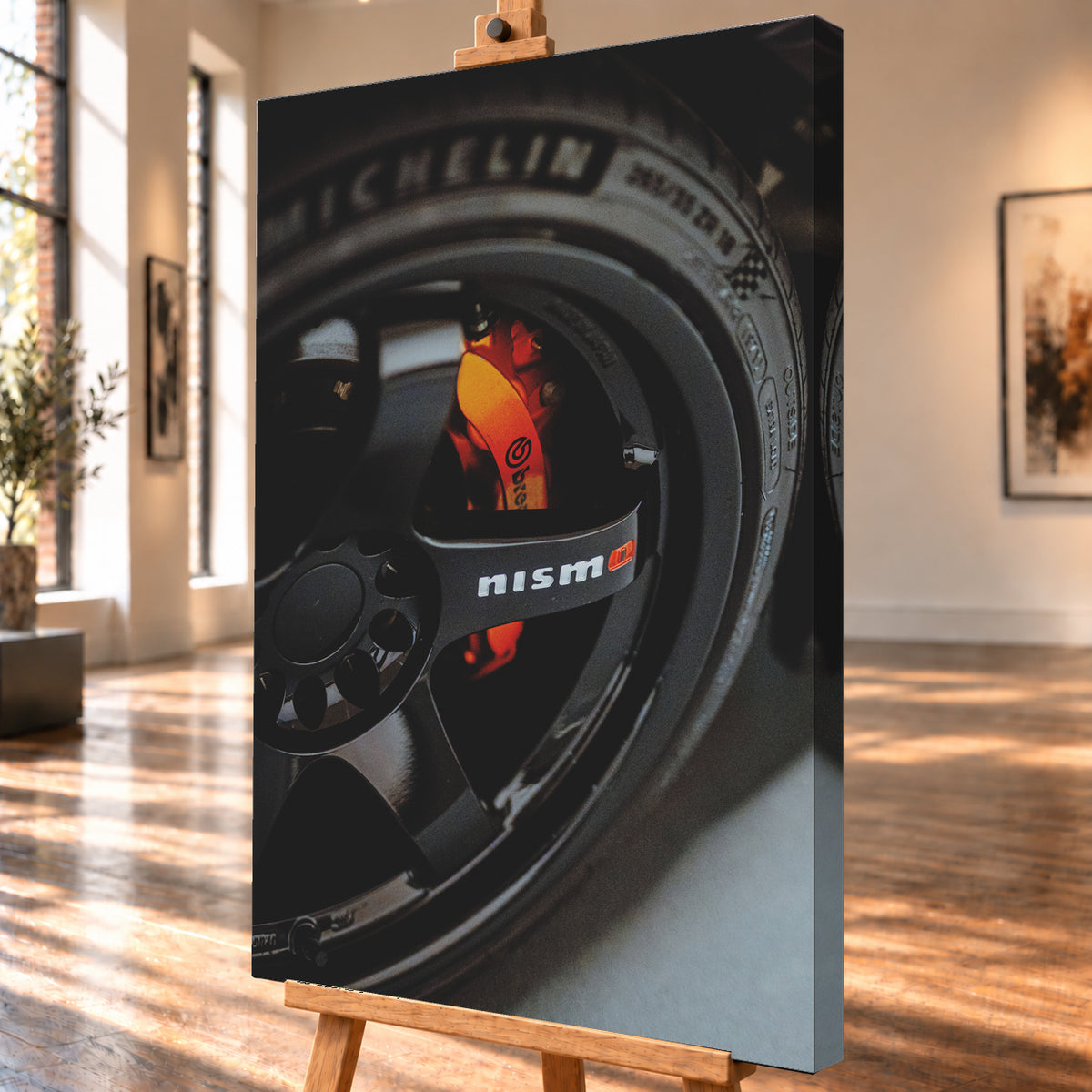 Nahaufnahme einer schwarzen Nismo-Mehrspeichenfelge mit orange-rotem Brembo-Bremssattel und dunkler Reifenflanke; Visualisierung als Leinwand-Wandbild, schräg von der Seite auf einer Holzstaffelei.