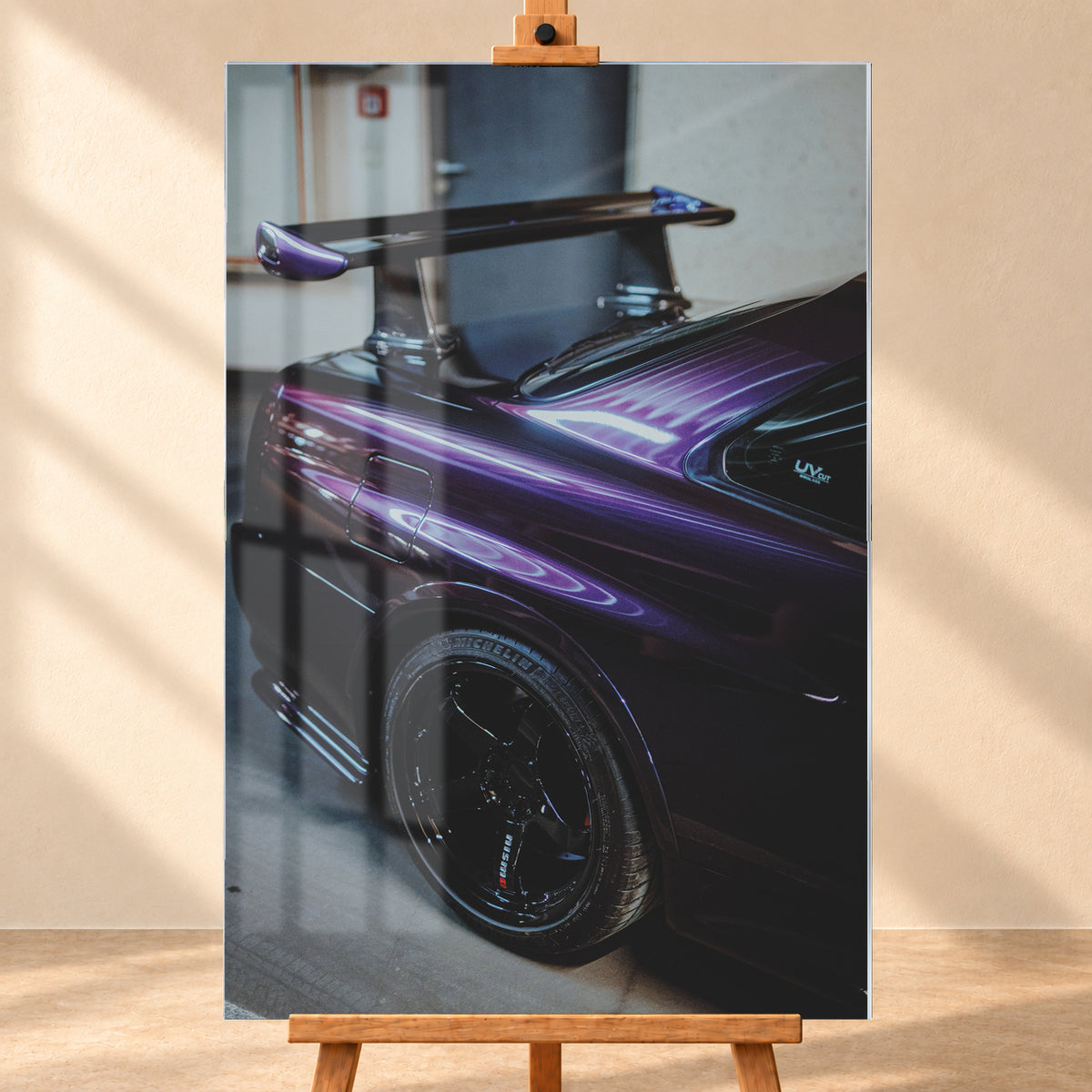 Nahaufnahme der hinteren Seitenpartie eines violetten Nissan Skyline R34 in einem Innenraum; sichtbar sind schwarzes Hinterrad, Radlauf, Heckscheibe und großer Heckflügel mit hellen Reflexionen; Visualisierung als Acrylglas-Wandbild, frontal auf einer Holzstaffelei.