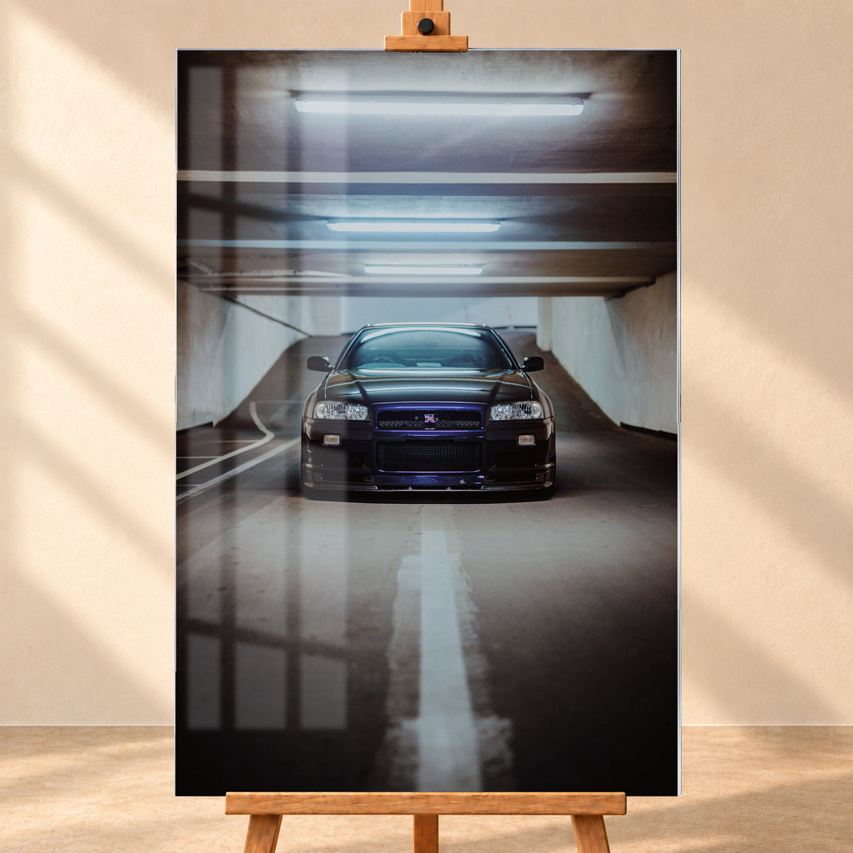 Dunkler Nissan Skyline R34 in frontaler Ansicht auf einer Betonrampe, unter kühlen weißen Deckenleuchten, mit mittiger Fahrbahnmarkierung und violettblauen Reflexen an Kühlergrill und Frontschürze; Visualisierung als Acrylglas-Wandbild, frontal auf einer Holzstaffelei.