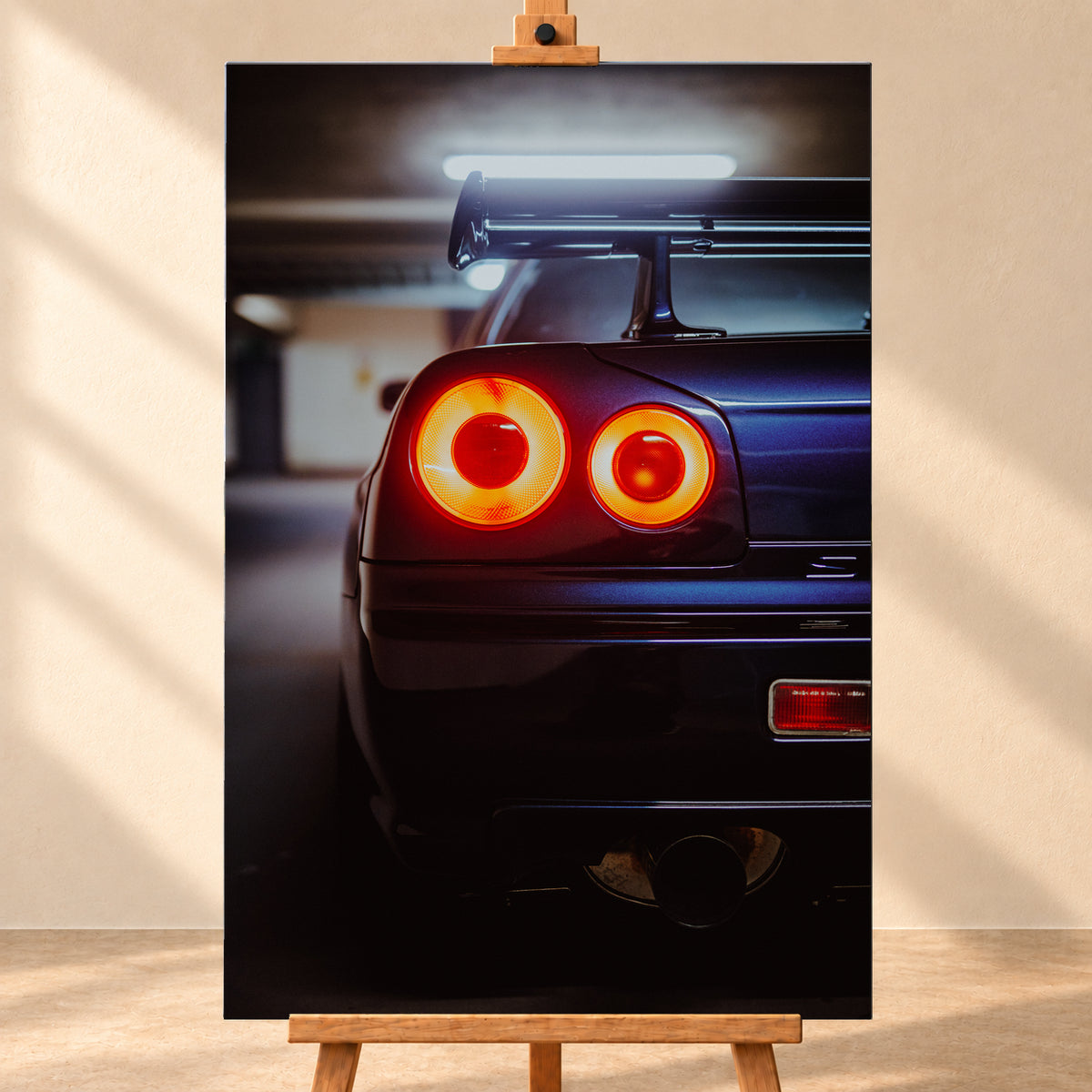 Nahaufnahme der Heckpartie eines dunklen Nissan Skyline R34 V-Spec mit zwei sichtbaren, rot-orange leuchtenden Rundrückleuchten, großem Heckflügel und rundem Auspuffendrohr vor unscharfem, kühl beleuchtetem Hintergrund; Visualisierung als Leinwand-Wandbild, frontal auf einer Holzstaffelei.