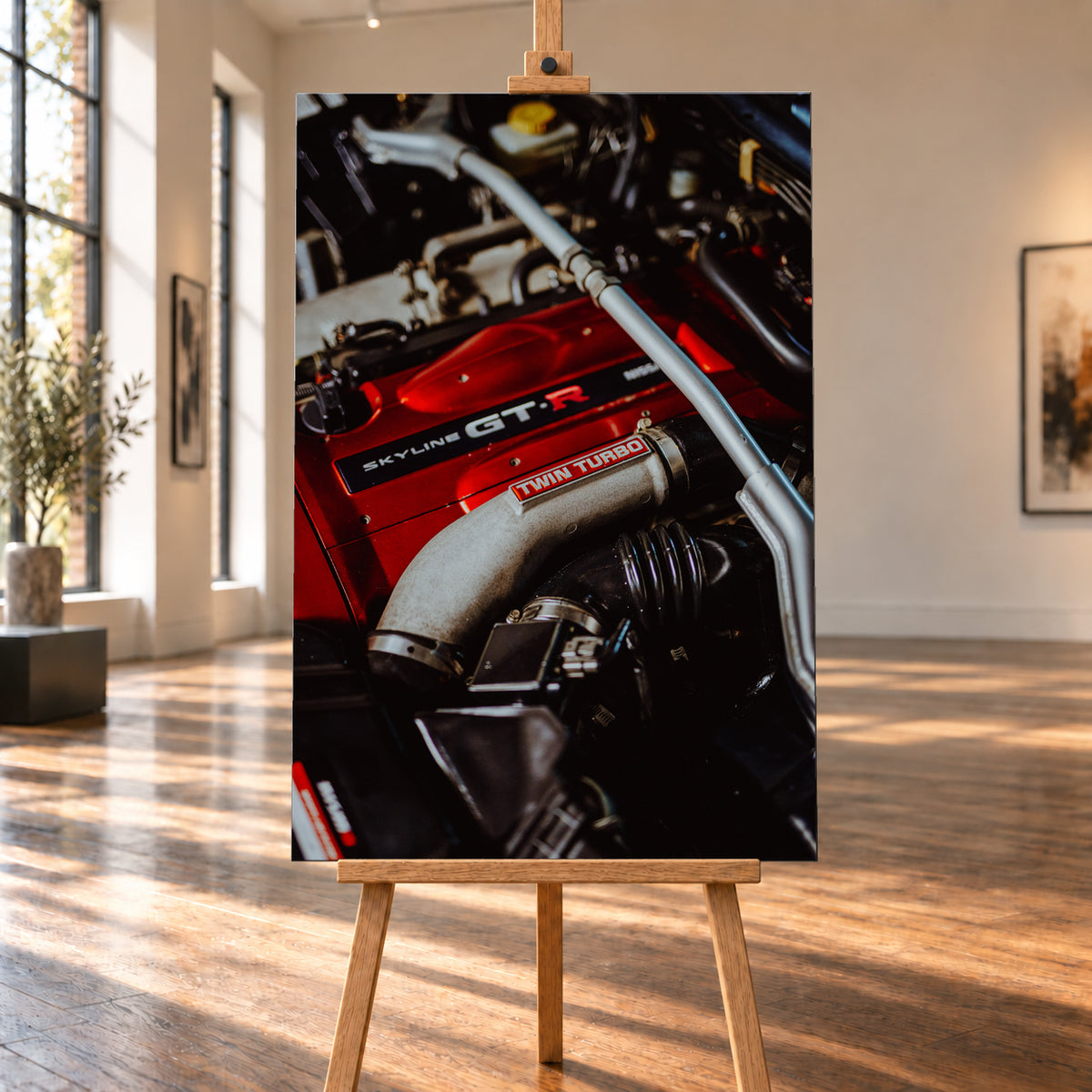Nahaufnahme eines Motorraums mit roter Abdeckung und sichtbarem SKYLINE GT-R-Schriftzug, hellem Rohr mit TWIN TURBO-Kennzeichnung, schwarzer Verrohrung und einer silbernen, diagonal verlaufenden Strebe; dunkler Hintergrund mit warmen Lichtreflexen; Visualisierung als Leinwand-Wandbild, auf einer Holzstaffelei in einem hellen Galerieraum.