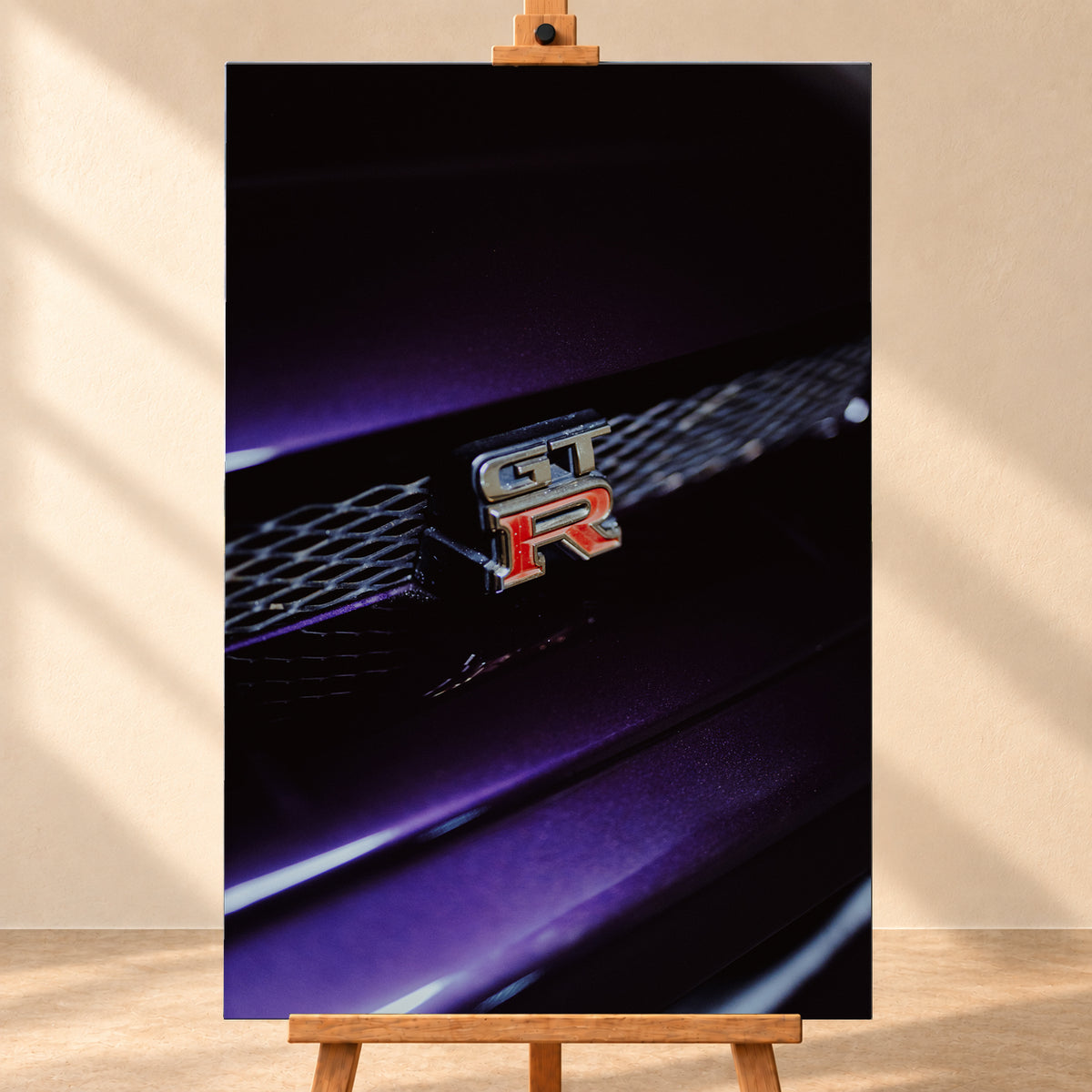 Nahaufnahme eines rot-silbernen GT-R-Emblems an einem dunklen Frontgrill, beleuchtet von violettem Licht und umgeben von tiefen Schatten; Visualisierung als Leinwand-Wandbild, frontal auf einer Holzstaffelei.