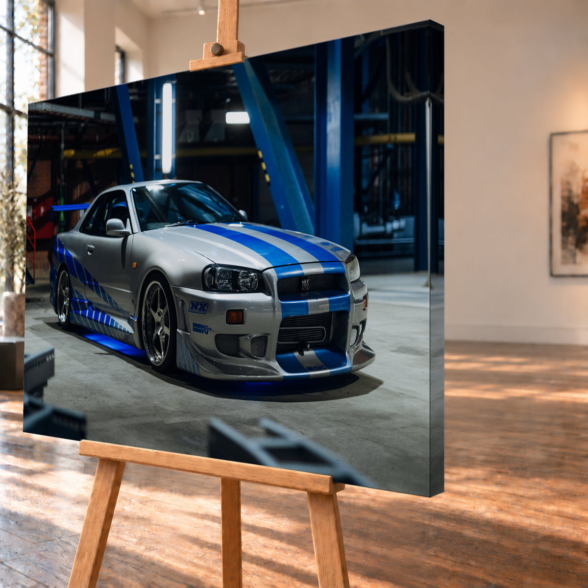 Silberner Nissan Skyline GT-R R34 mit blauen Streifen und Seitengrafik in tiefer Front-Dreiviertelansicht. Das Fahrzeug steht auf Beton in einer dunklen Industriehalle mit blauen Stahlträgern, hellen Leuchten und blauem Licht unter der Karosserie. Frontschürze und großer Heckflügel sind sichtbar; Visualisierung als Leinwand-Wandbild, schräg von der Seite auf einer Holzstaffelei.
