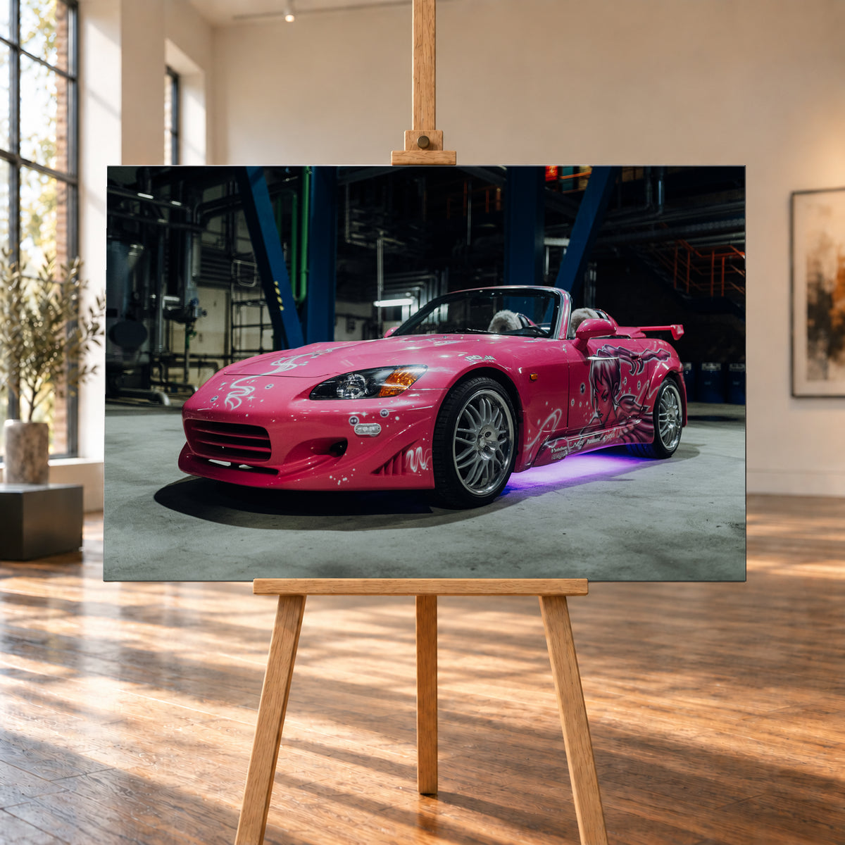 Pinker Honda S2000 als offener Roadster in Front-Dreiviertelansicht auf einem Betonboden in einer dunklen Industriehalle; sichtbar sind weiße Dekorgrafiken, eine große Frauenfigur an der Seite, silberne Mehrspeichenräder, ein Heckflügel und violett-blaues Licht unter dem Fahrzeug; Visualisierung als Leinwand-Wandbild, auf einer Holzstaffelei in einem hellen Galerieraum.
