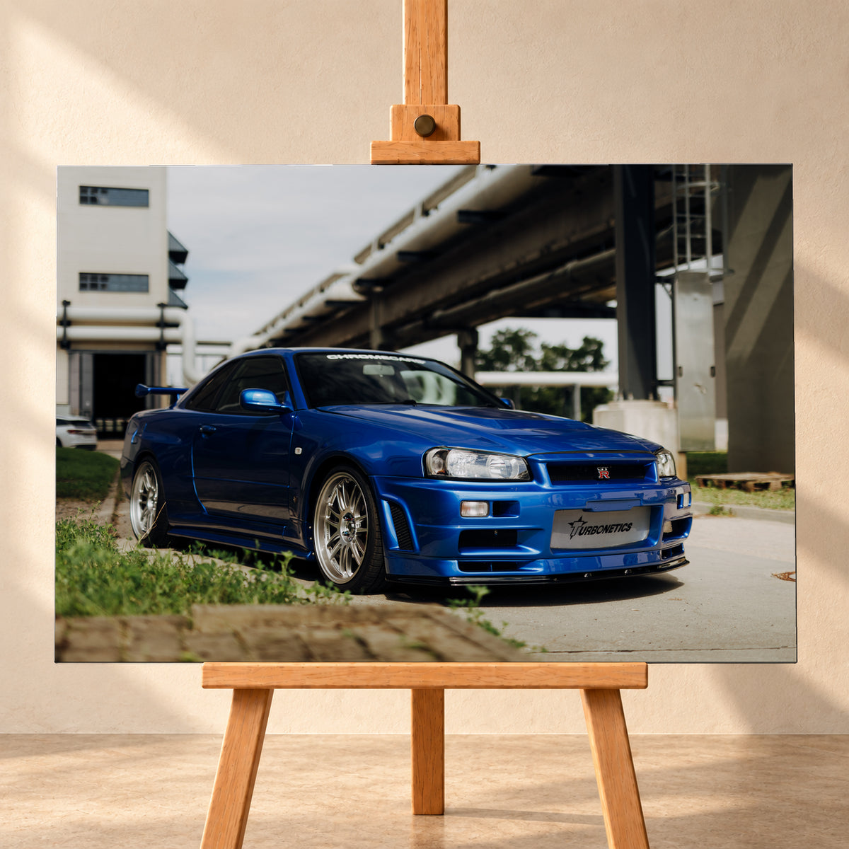 Blauer Nissan Skyline R34 GT-R in tiefer Front-Dreiviertelansicht auf Asphalt unter einer erhöhten Betonstruktur; sichtbar sind silberne Mehrspeichenräder, Heckflügel, GT-R-Emblem und Gras am Bildrand; Visualisierung als Leinwand-Wandbild, frontal auf einer Holzstaffelei.