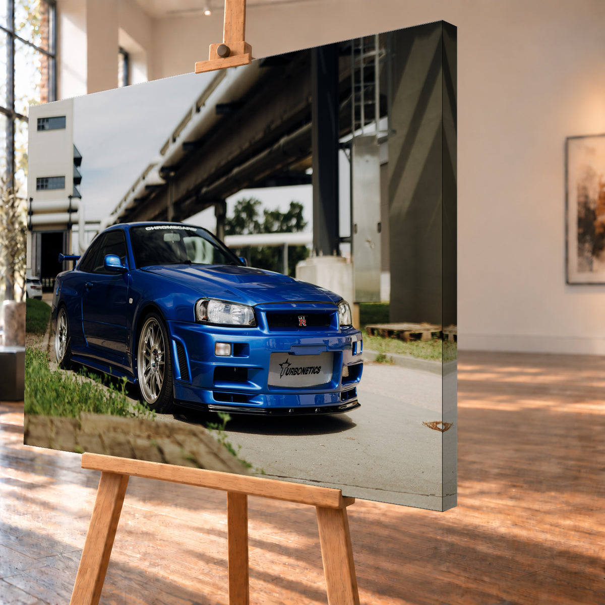 Blauer Nissan Skyline R34 GT-R in tiefer Front-Dreiviertelansicht auf Asphalt unter einer erhöhten Betonstruktur; sichtbar sind silberne Mehrspeichenräder, Heckflügel, GT-R-Emblem und Gras am Bildrand; Visualisierung als Leinwand-Wandbild, schräg von der Seite auf einer Holzstaffelei.