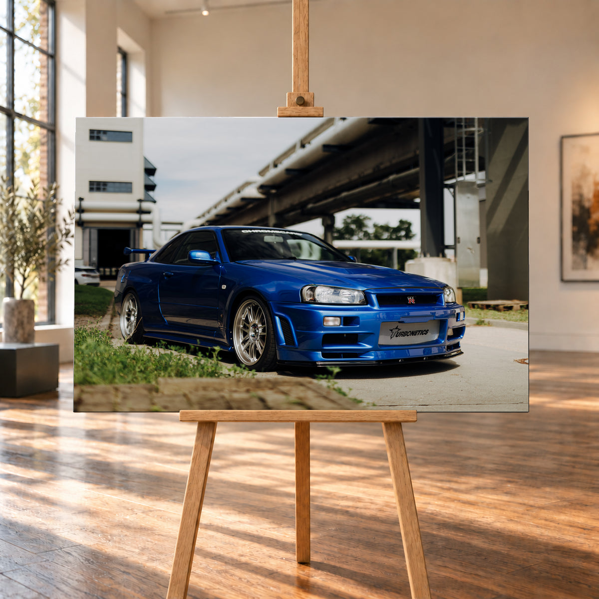 Blauer Nissan Skyline R34 GT-R in tiefer Front-Dreiviertelansicht auf Asphalt unter einer erhöhten Betonstruktur; sichtbar sind silberne Mehrspeichenräder, Heckflügel, GT-R-Emblem und Gras am Bildrand; Visualisierung als Leinwand-Wandbild, auf einer Holzstaffelei in einem hellen Galerieraum.