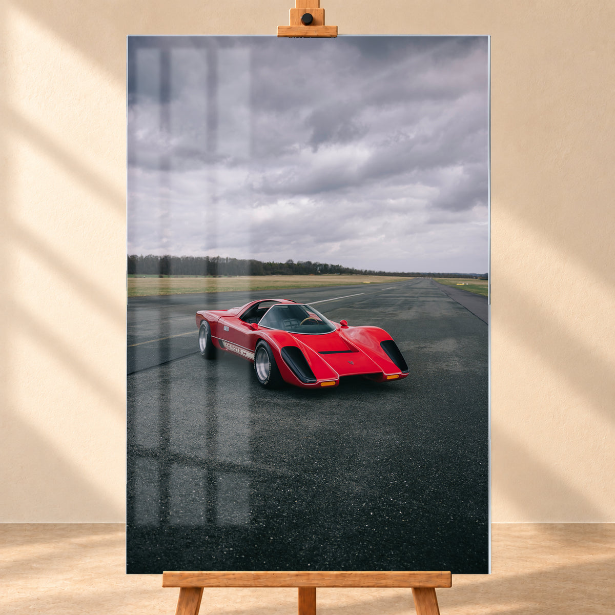 Roter Coyote X in Front-Dreiviertelansicht auf einer breiten dunklen Asphaltbahn. Das hochformatige Bild zeigt flache Wiesen, einen niedrigen Baumhorizont und einen ausgedehnten grauen Wolkenhimmel; Personen sind nicht sichtbar; Visualisierung als Acrylglas-Wandbild, frontal auf einer Holzstaffelei.