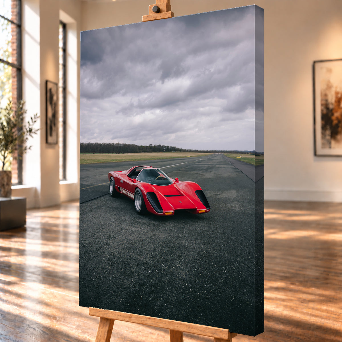 Roter Coyote X in Front-Dreiviertelansicht auf einer breiten dunklen Asphaltbahn. Das hochformatige Bild zeigt flache Wiesen, einen niedrigen Baumhorizont und einen ausgedehnten grauen Wolkenhimmel; Personen sind nicht sichtbar; Visualisierung als Leinwand-Wandbild, schräg von der Seite auf einer Holzstaffelei.