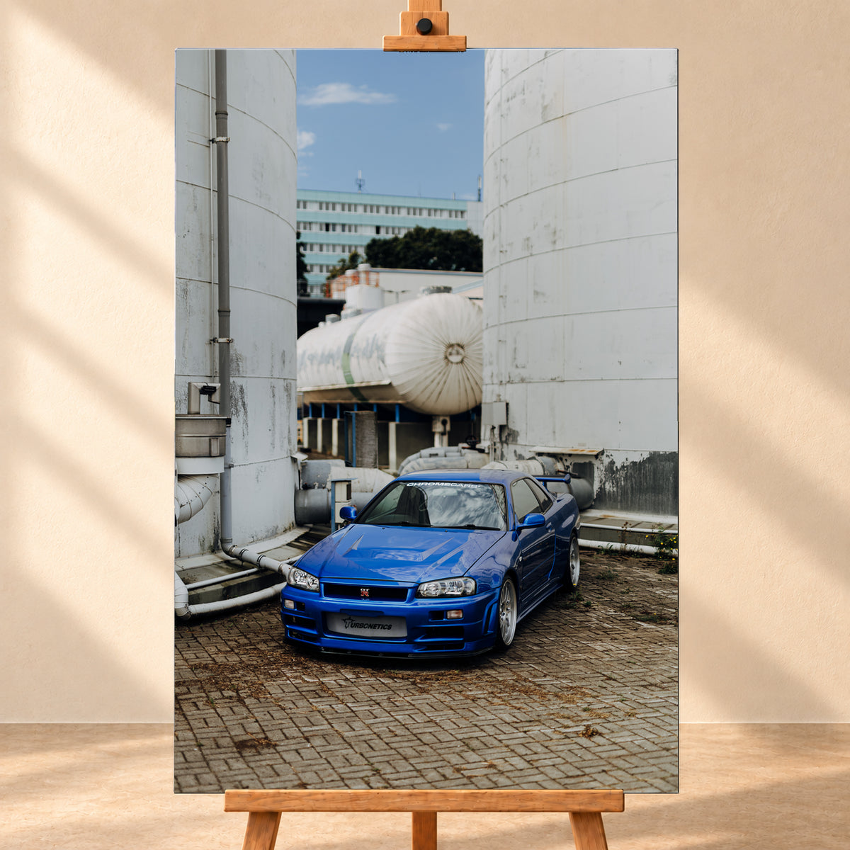 Blauer Nissan Skyline R34 GT-R in frontaler Dreiviertelansicht auf gepflastertem Industriegelände zwischen hohen weißen Behältern, Tanks und Rohrleitungen bei Tageslicht; Visualisierung als Leinwand-Wandbild, frontal auf einer Holzstaffelei.