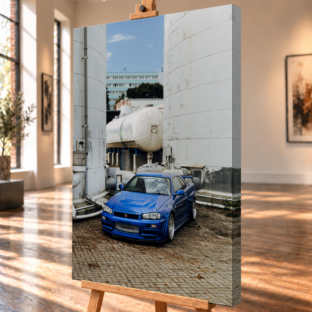 Blauer Nissan Skyline R34 GT-R in frontaler Dreiviertelansicht auf gepflastertem Industriegelände zwischen hohen weißen Behältern, Tanks und Rohrleitungen bei Tageslicht; Visualisierung als Leinwand-Wandbild, schräg von der Seite auf einer Holzstaffelei.
