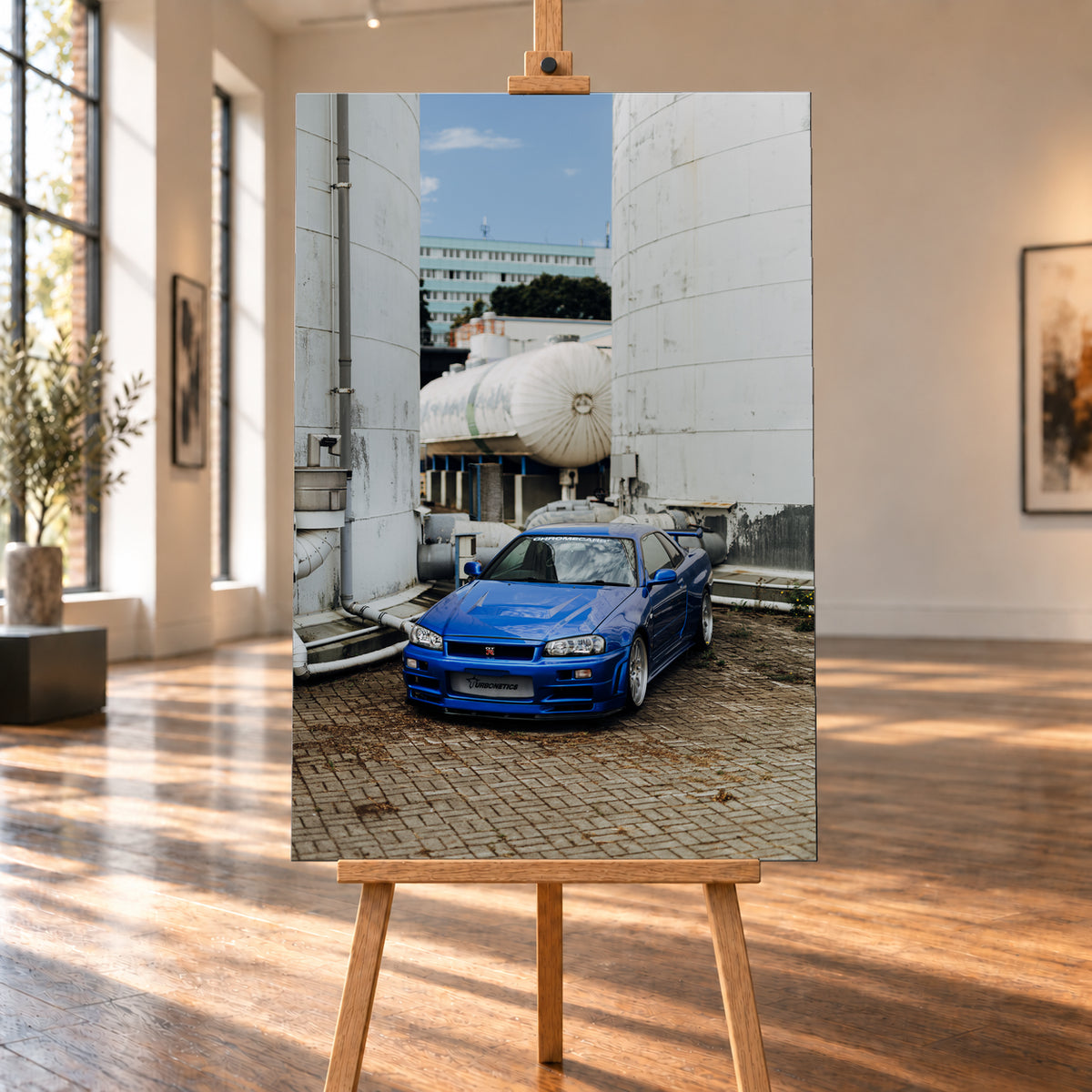 Blauer Nissan Skyline R34 GT-R in frontaler Dreiviertelansicht auf gepflastertem Industriegelände zwischen hohen weißen Behältern, Tanks und Rohrleitungen bei Tageslicht; Visualisierung als Leinwand-Wandbild, auf einer Holzstaffelei in einem hellen Galerieraum.