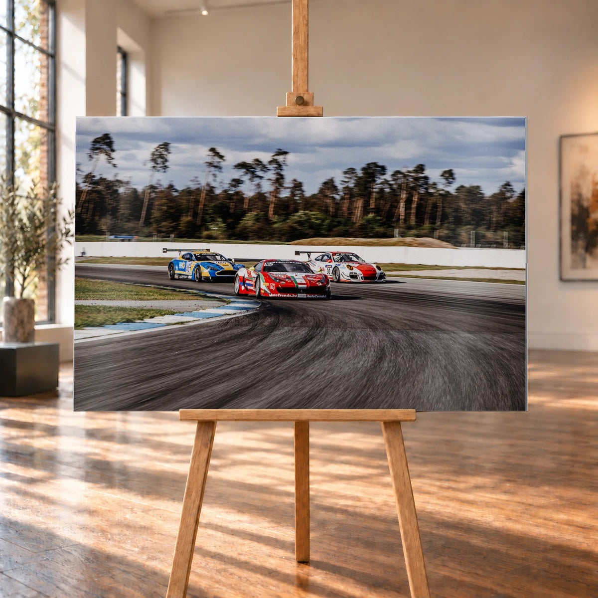 Aston Martin, Porsche und Ferrari GT3 fahren dicht gestaffelt durch eine Kurve; die Mehrwagen-Szene zeigt echtes Renngeschehen. Motiv „Racing Echo“ als Alu gebürstet-Wandbild, auf einer Holzstaffelei in einem hellen Galerieraum.
