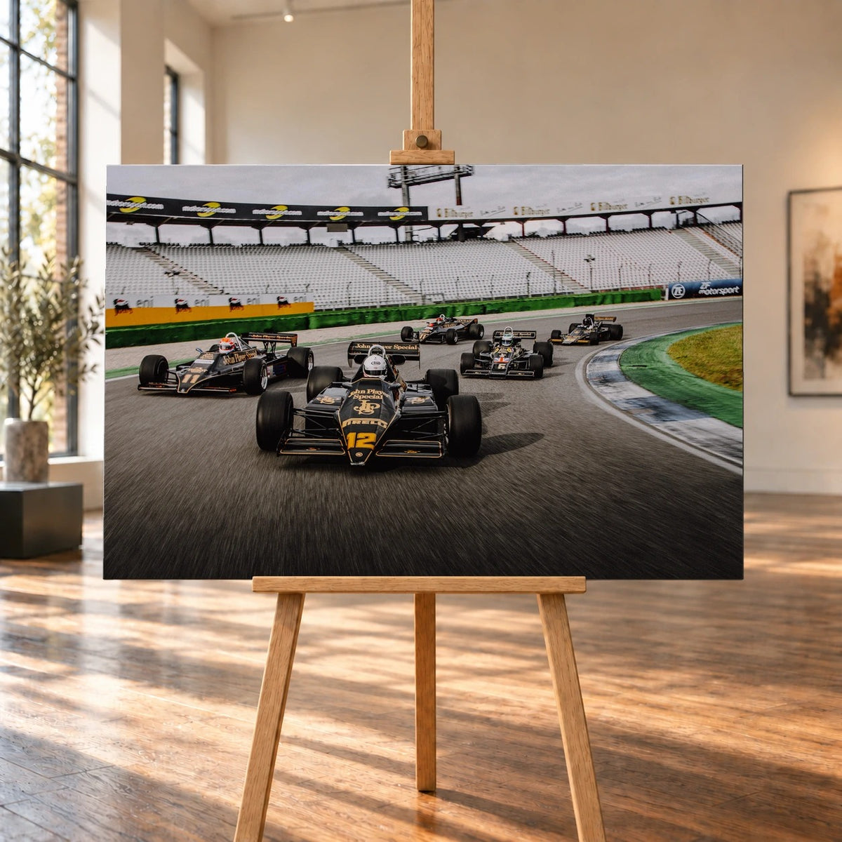 Raumvisualisierung eines querformatigen Leinwandbilds auf einer Holzstaffelei in einem hellen Raum mit großen Fenstern und Holzboden. Das Bild zeigt fünf schwarz-goldene Lotus-Formelrennwagen in einer Kurve vor leeren Tribünen.