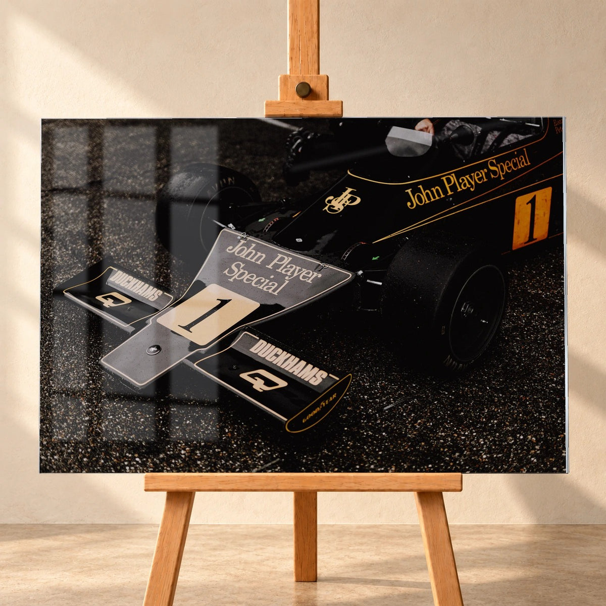 Frontal visualisiertes Acrylglasbild auf einer Holzstaffelei vor heller Wand, mit Fensterreflexionen auf der Bildfläche. Zu sehen ist die Front des schwarz-goldenen Lotus 76 mit Startnummer 1 und „John Player Special“-Schriftzug.