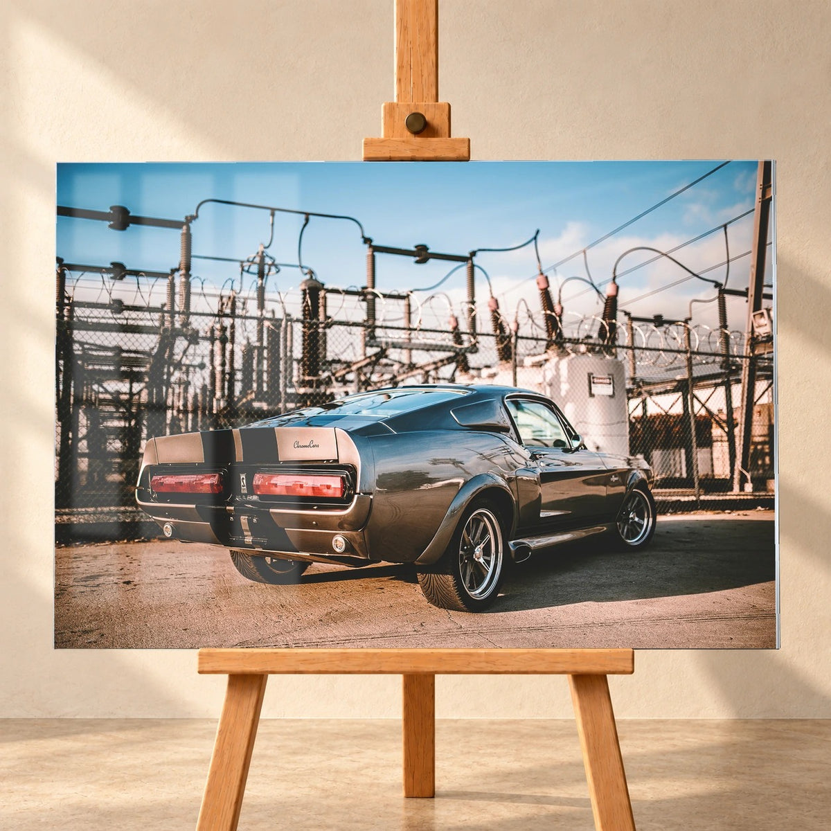 Frontal visualisiertes Acrylglas-Wandbild auf einer Holzstaffelei vor heller Wand, mit sichtbaren Fensterreflexionen. Das Motiv zeigt Eleanor, einen grauen Ford Mustang Shelby GT500 mit schwarzen Streifen, schräg von hinten vor einem Umspannwerk.