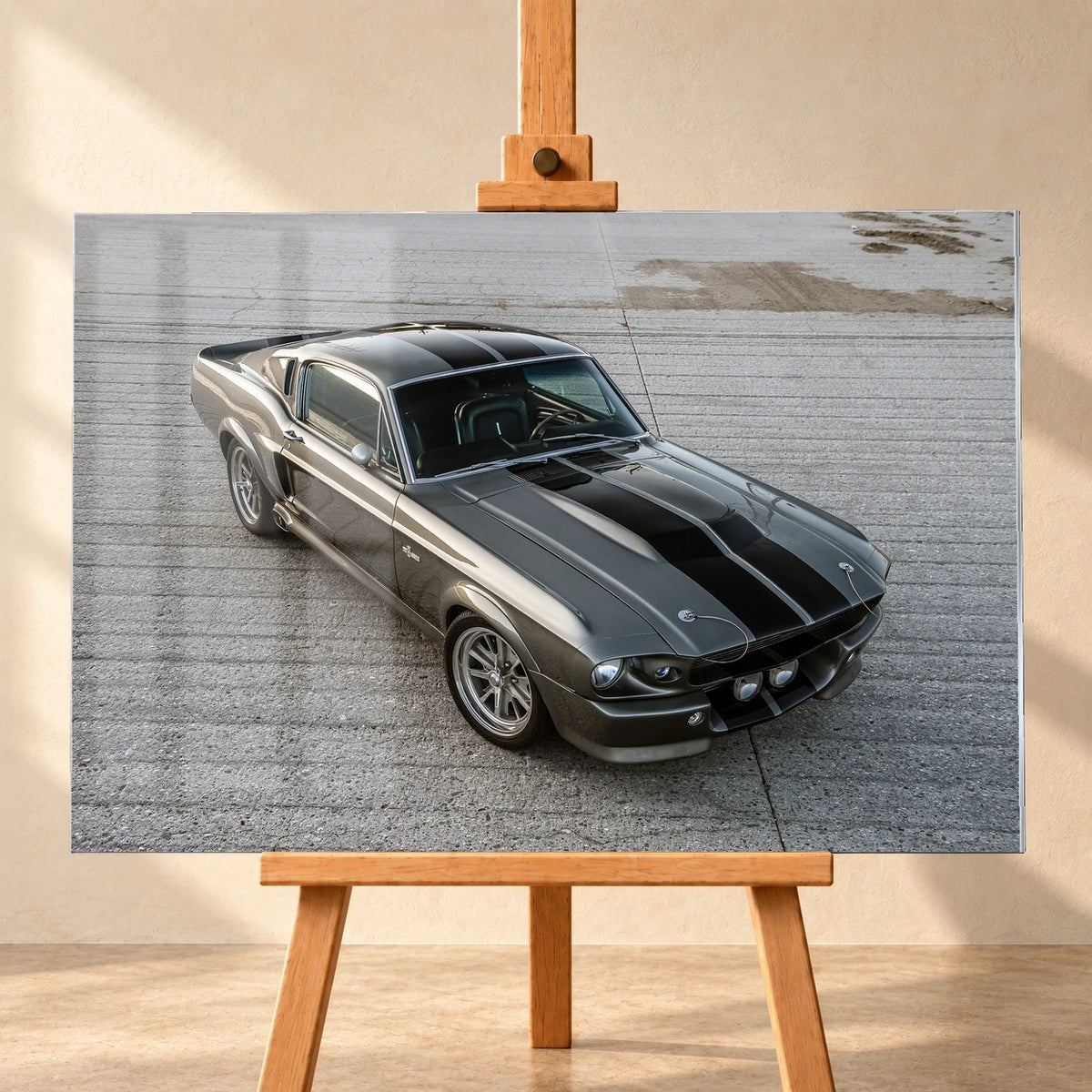 Frontalansicht eines querformatigen Acrylglasbilds als Visualisierung auf einer Holzstaffelei vor heller Wand. Abgebildet ist Eleanor, der graue Ford Mustang Shelby GT500 mit schwarzen Doppelstreifen, aus erhöhter Perspektive schräg von vorn auf einer Betonfläche.