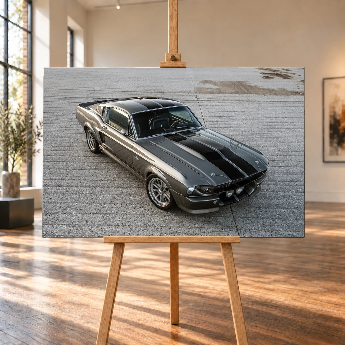Raumvisualisierung eines Leinwandbilds im Querformat auf einer Holzstaffelei in einem hellen Galerieraum mit großen Fenstern und Holzboden. Das Motiv zeigt den grauen Ford Mustang Shelby GT500 Eleanor mit schwarzen Doppelstreifen schräg von vorn und oben auf Beton.