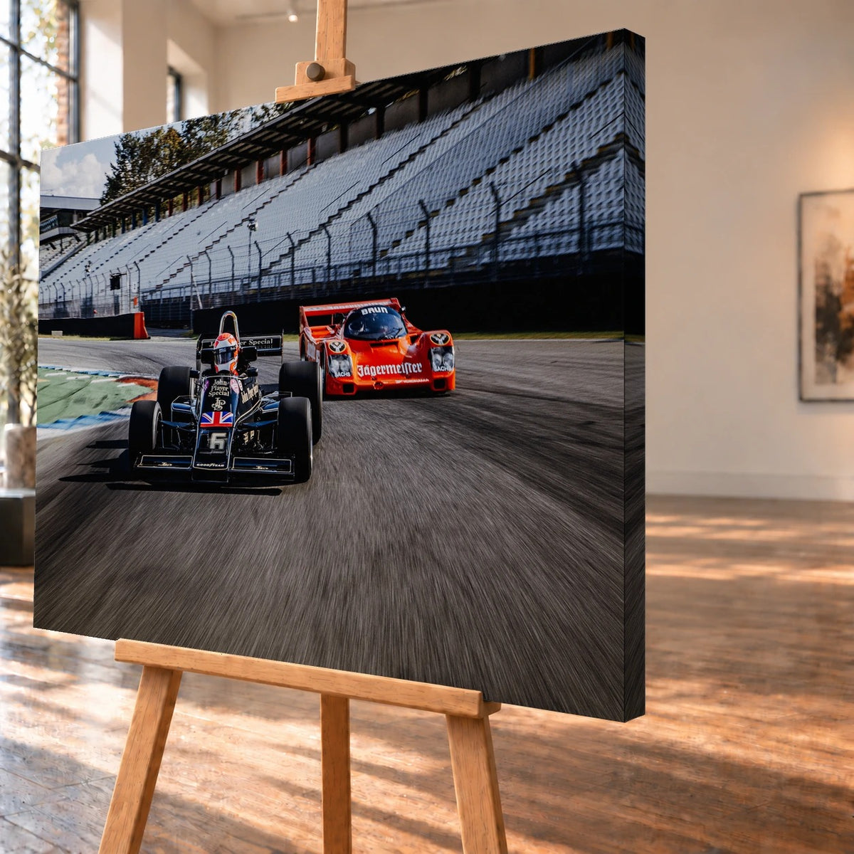 Schrägansicht eines Leinwandbilds als Visualisierung auf einer Holzstaffelei; die rechte, mit dem Motiv bedruckte Seitenkante ist sichtbar. Ein schwarzer Lotus 77 fährt vor einem orangefarbenen Porsche 962C in Brun/Jägermeister-Lackierung durch eine Rennstreckenkurve vor leeren Tribünen.