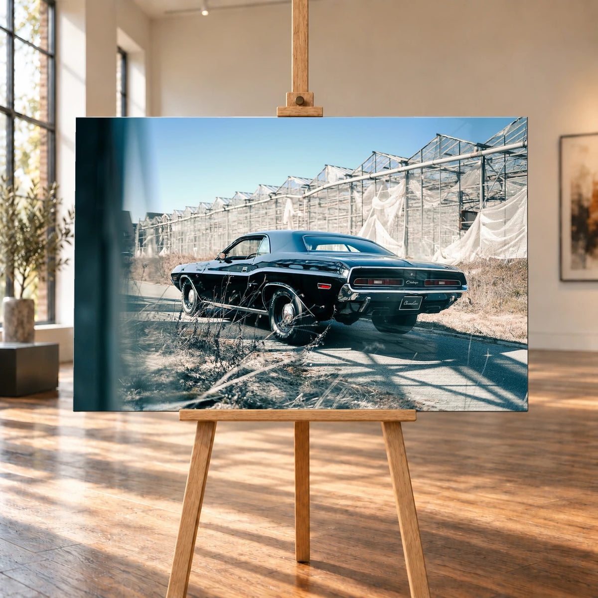 Raumvisualisierung mit querformatigem Leinwandbild auf einer Holzstaffelei in einem hellen Raum mit hohen Fenstern und Holzboden. Auf dem Bild ist ein schwarzer Dodge Challenger 426 Hemi (1971) schräg von hinten vor einer langen Gewächshausreihe zu sehen.