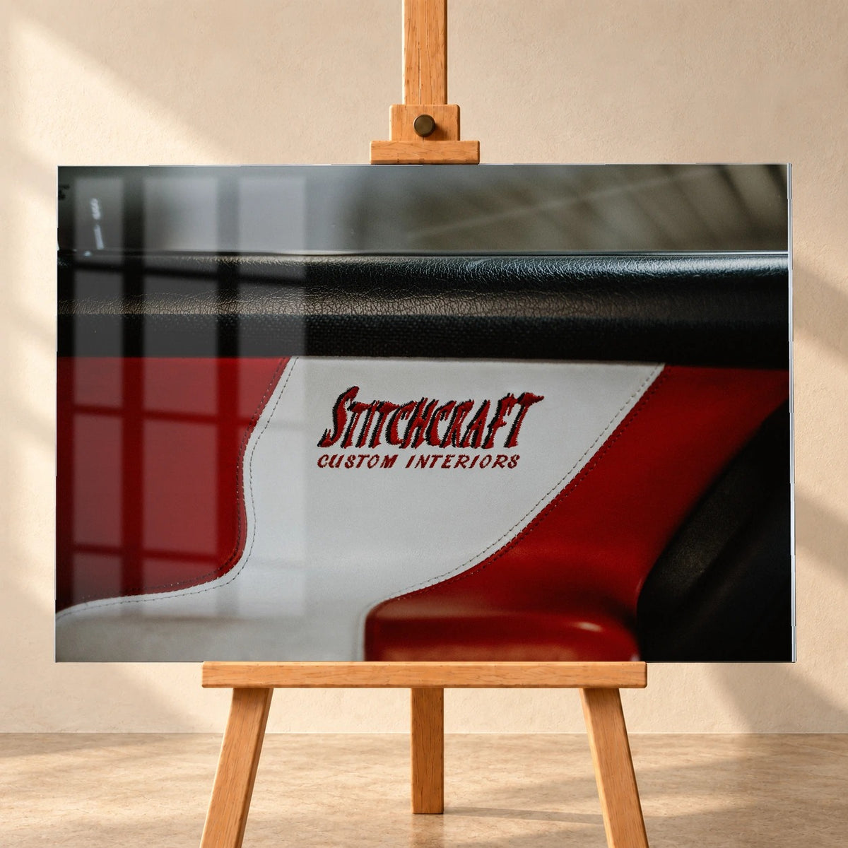 Visualisiertes Acrylglasbild in frontaler Ansicht auf einer Holzstaffelei. Eine Fensterspiegelung liegt über dem Motiv der rot-weiß-schwarzen VW-Jetta-Türverkleidung mit der Stickerei „Stitchcraft Custom Interiors“ auf dem weißen Einsatz.