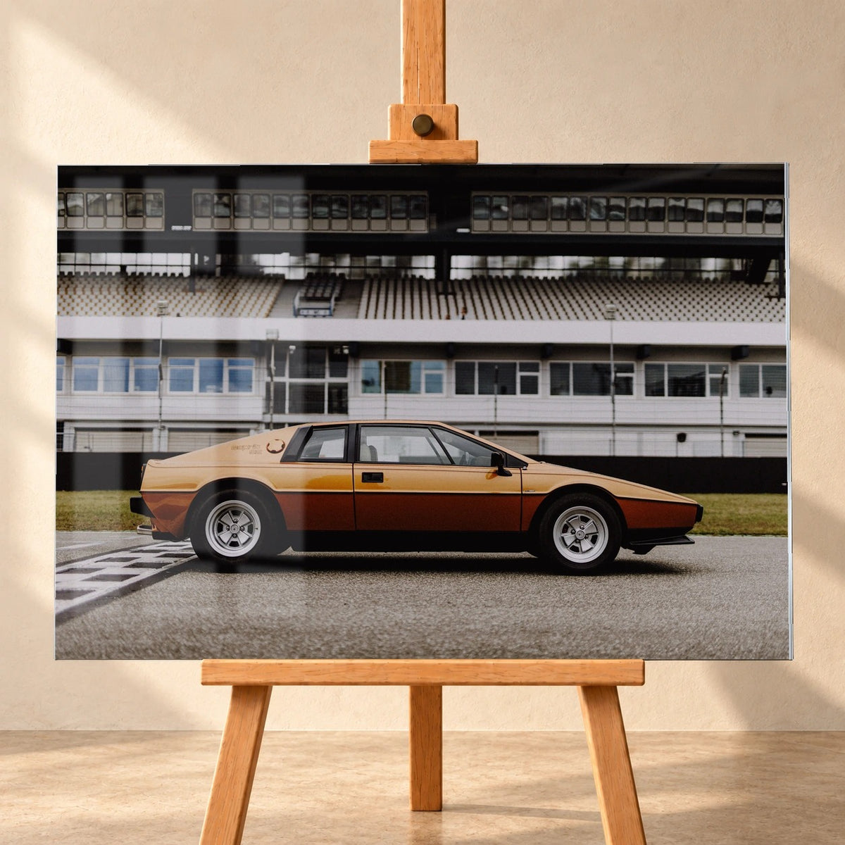 Visualisierung eines Acrylglasbilds in frontaler Ansicht auf einer Holzstaffelei. Fensterreflexe liegen über dem Motiv des gold- und kupferfarbenen Lotus Esprit Gold Edition in Seitenansicht vor einer Rennstreckentribüne.