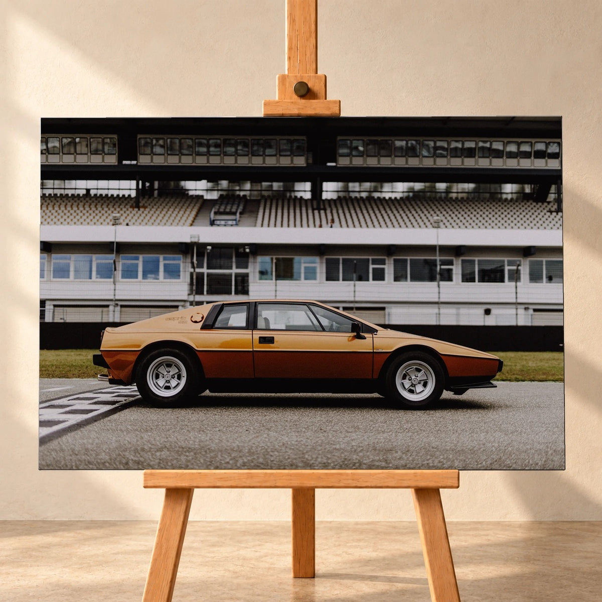 Frontale Visualisierung eines Leinwandbilds auf einer Holzstaffelei vor heller Wand. Das Motiv zeigt den gold- und kupferfarbenen Lotus Esprit Gold Edition in Seitenansicht vor einer Rennstreckentribüne.