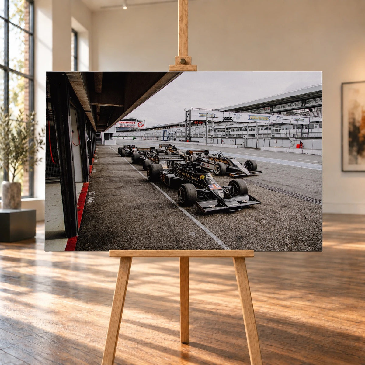 Fünf schwarz-goldene Lotus-Rennwagen in der Boxengasse als Motiv eines querformatigen Leinwandbilds. Die Raumvisualisierung zeigt das Bild frontal auf einer Holzstaffelei in einem hellen Raum mit hohen Fenstern und Holzboden.