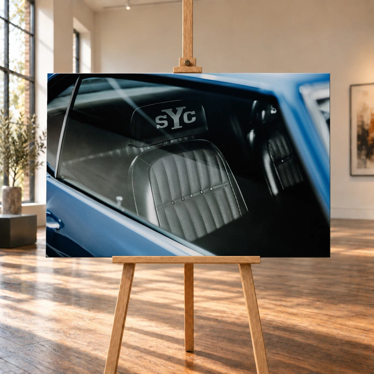 Raumvisualisierung eines querformatigen Leinwandbildes auf einer Holzstaffelei in einem hellen Raum mit großen Fenstern und Holzboden. Das Motiv zeigt den Blick in den blauen Chevrolet Camaro Yenko, im Mittelpunkt den Vordersitz mit „sYc“-Kopfstütze.