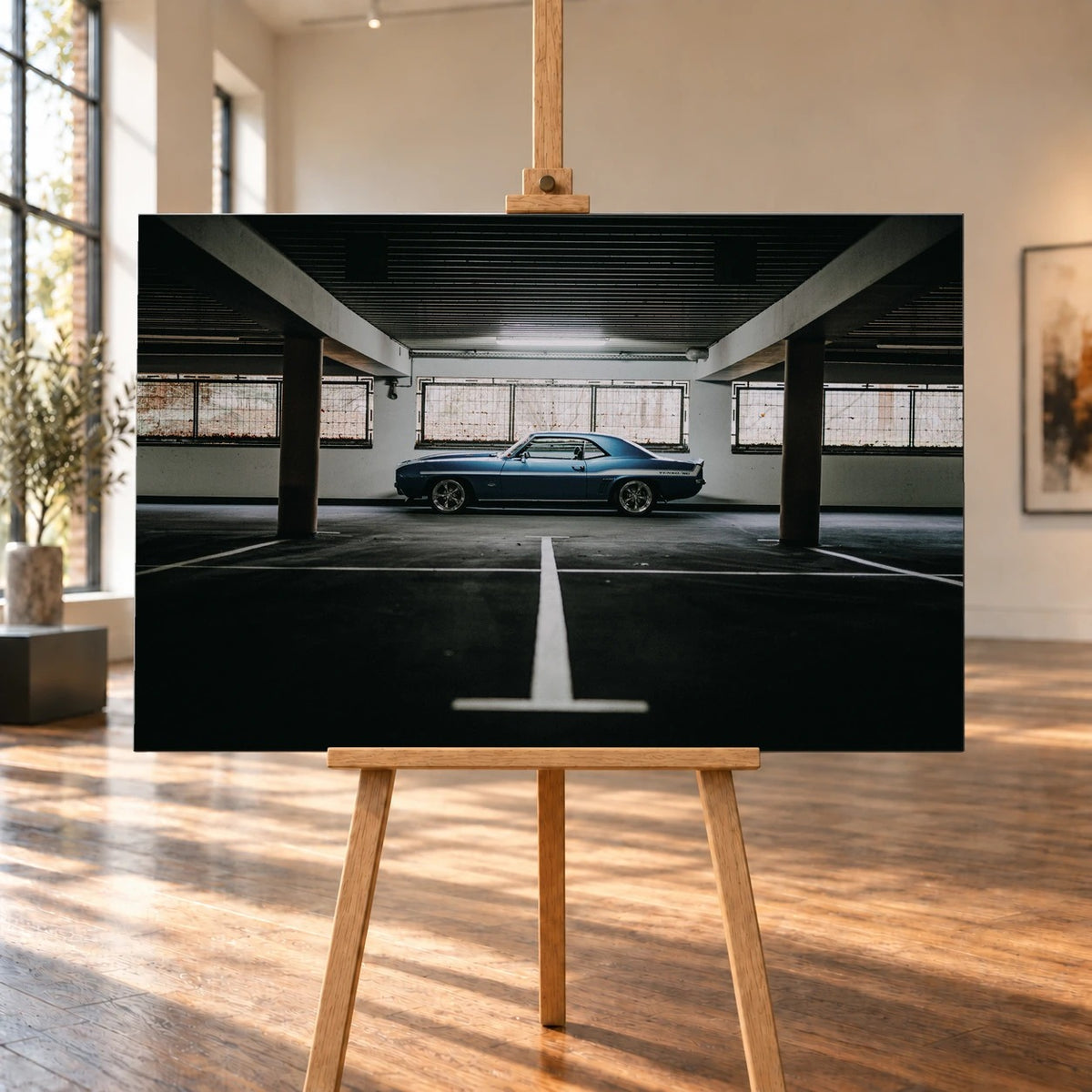 Ein querformatiges Leinwandbild ist auf einer Holzstaffelei in einem hellen Raum mit großen Fenstern und Holzboden visualisiert. Im Bild steht ein blauer Chevrolet Camaro Yenko seitlich zwischen den Säulen einer Garage.