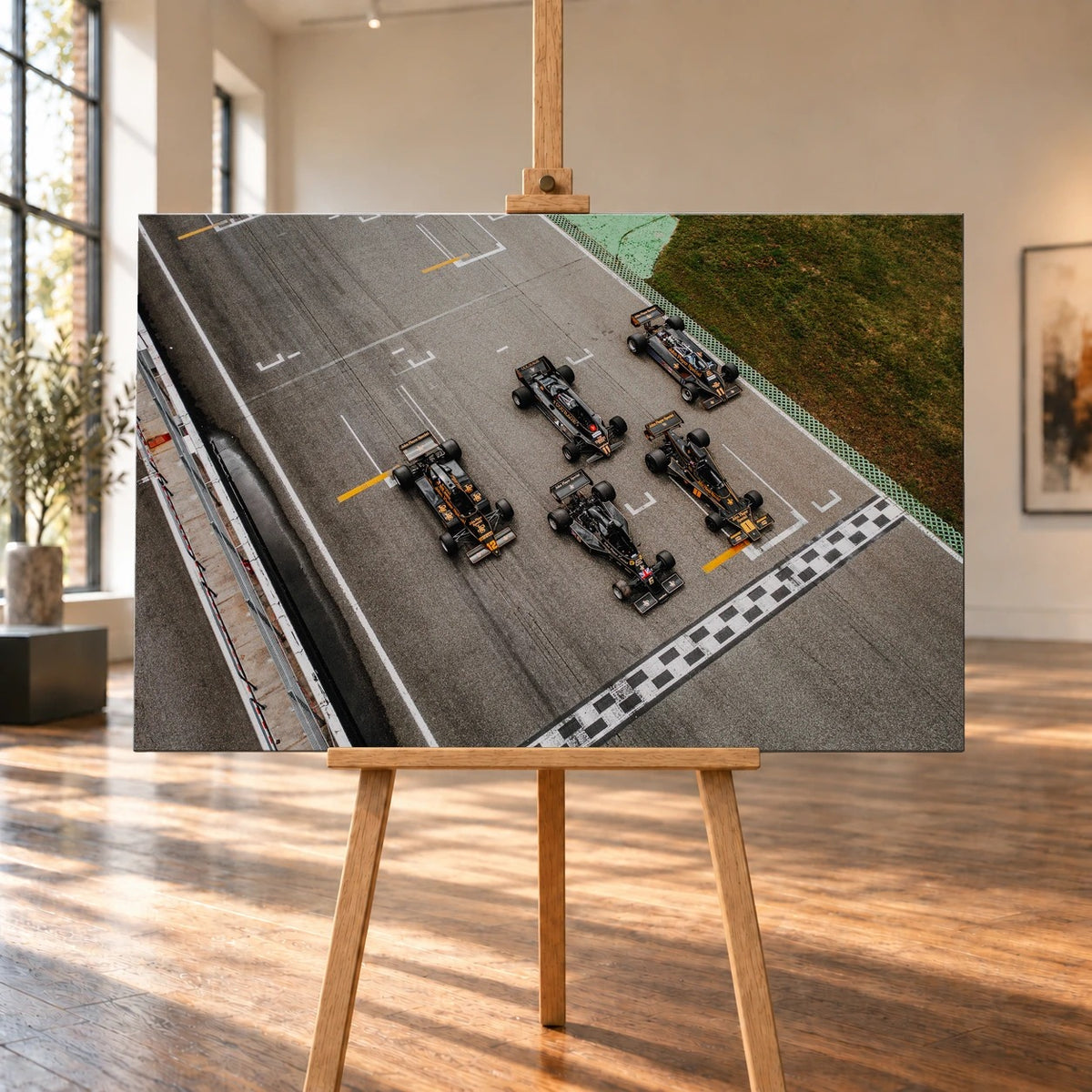 Raumvisualisierung eines querformatigen Leinwandbilds auf einer Holzstaffelei in einem hellen Raum mit großen Fenstern und Holzboden. Auf dem Bild sind fünf schwarz-goldene Lotus-Rennwagen von oben vor der karierten Start-Ziel-Linie zu sehen.