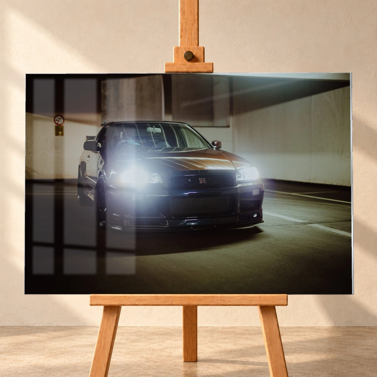 Visualisierung eines Acrylglasbilds auf einer Holzstaffelei, frontal vor heller Wand. Das querformatige Motiv zeigt einen Nissan Skyline GT-R R34 mit hellen Scheinwerfern im Parkhaus. Auf der dargestellten Bildoberfläche sind Fensterspiegelungen sichtbar.
