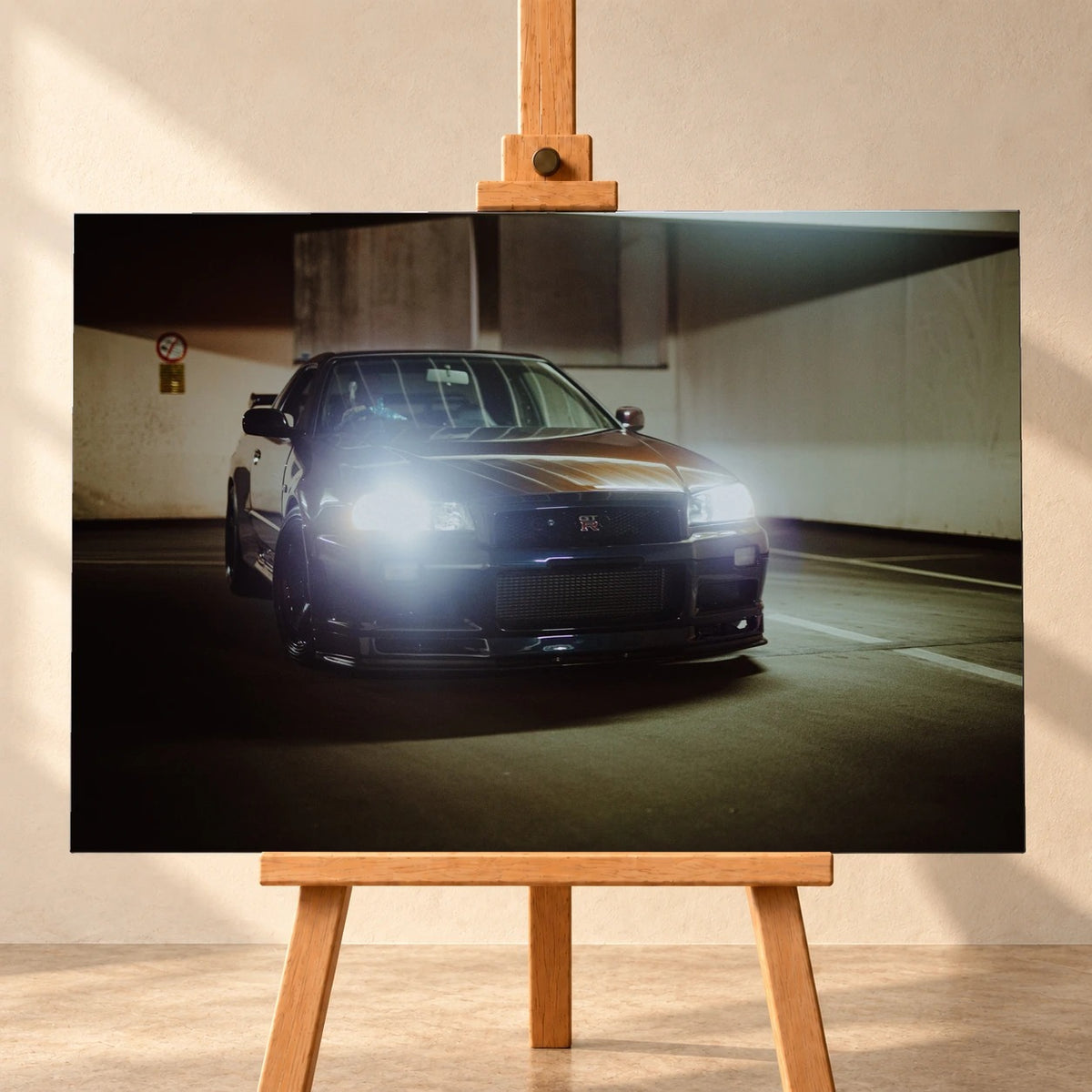 Frontale Visualisierung eines Leinwandbilds im Querformat auf einer Holzstaffelei vor heller Wand. Das Motiv zeigt einen dunklen Nissan Skyline GT-R R34 mit leuchtenden Scheinwerfern in schräger Frontansicht im Parkhaus.