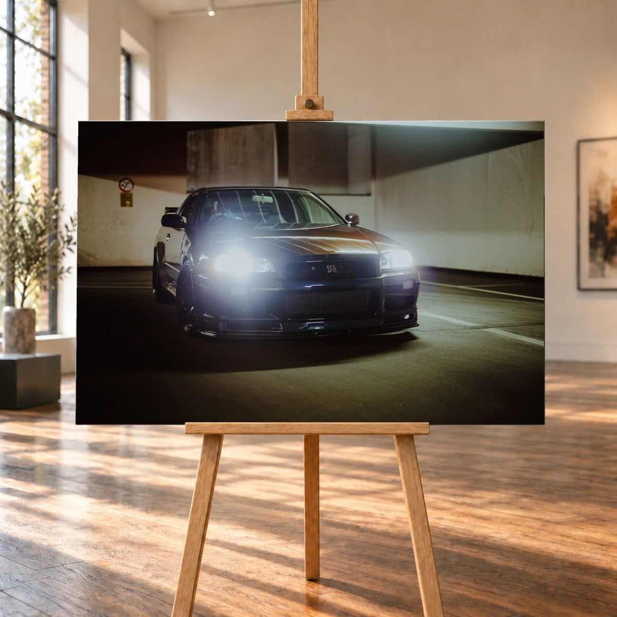 Raumvisualisierung eines querformatigen Leinwandbilds auf einer Holzstaffelei in einem hellen Raum mit großen Fenstern. Abgebildet ist ein dunkler Nissan Skyline GT-R R34 im Parkhaus, dessen Scheinwerfer zur Kamera leuchten.