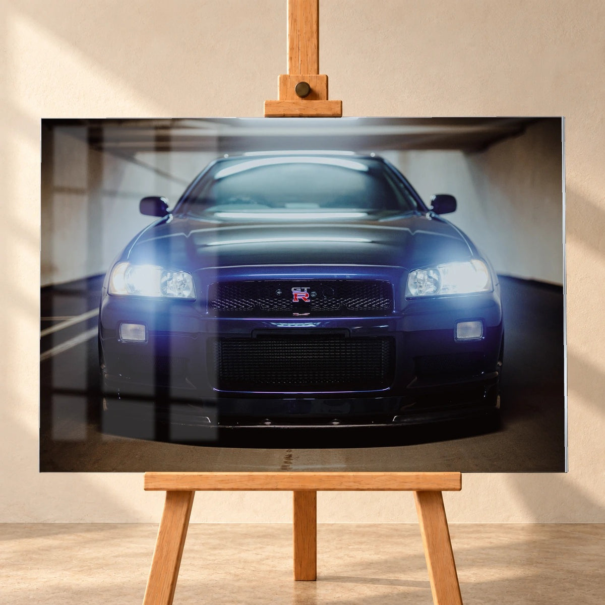 Visualisierung eines Acrylglas-Wandbilds auf einer Holzstaffelei vor heller Wand, mit sichtbaren Fensterspiegelungen auf der Bildfläche. Das Motiv zeigt die dunkelblau-violette Front eines Nissan Skyline GT-R R34 mit leuchtenden Scheinwerfern.