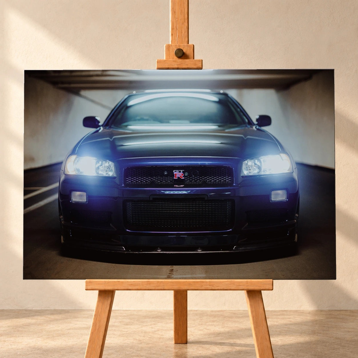 Visualisierung eines querformatigen Leinwandbilds auf einer Holzstaffelei vor heller Wand. Das Motiv zeigt einen dunkelblau-violetten Nissan Skyline GT-R R34 frontal in einem garagenartigen Innenraum, mit leuchtenden Scheinwerfern.
