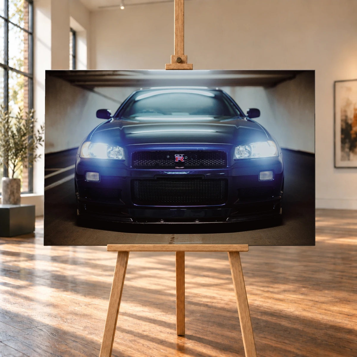 Raumvisualisierung eines Leinwandbilds auf einer Holzstaffelei in einem hellen Raum mit hohen Fenstern und Holzboden. Das querformatige Motiv zeigt einen Nissan Skyline GT-R R34 frontal mit leuchtenden Scheinwerfern.