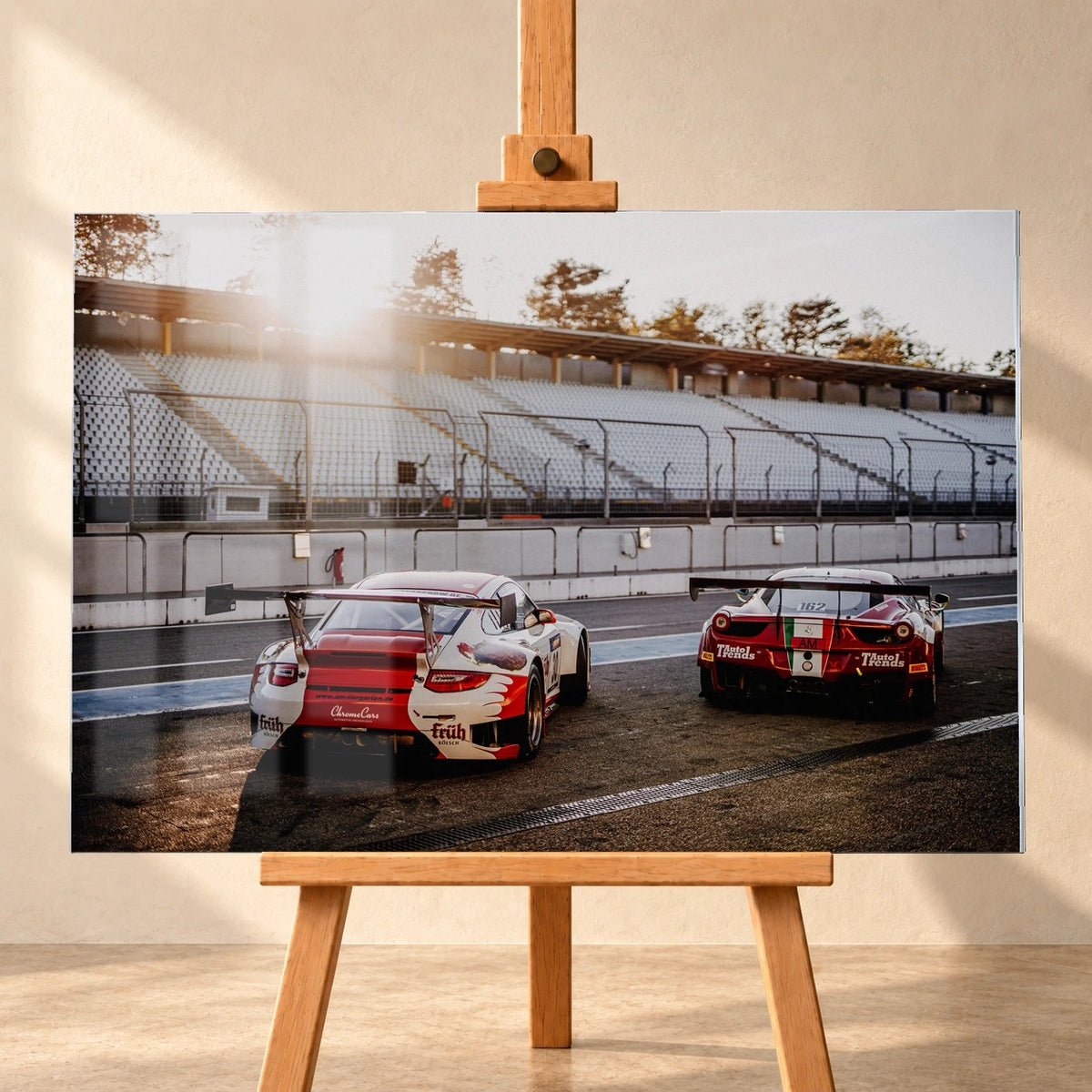 Visualisierung eines Acrylglasbilds im Querformat, frontal auf einer Holzstaffelei vor heller Wand präsentiert. Sichtbare Spiegelungen liegen über dem Motiv mit einem rot-weißen Porsche 911 GT3 R und einem roten Ferrari 458 GT3, von hinten in der Boxengasse vor leeren Tribünen.