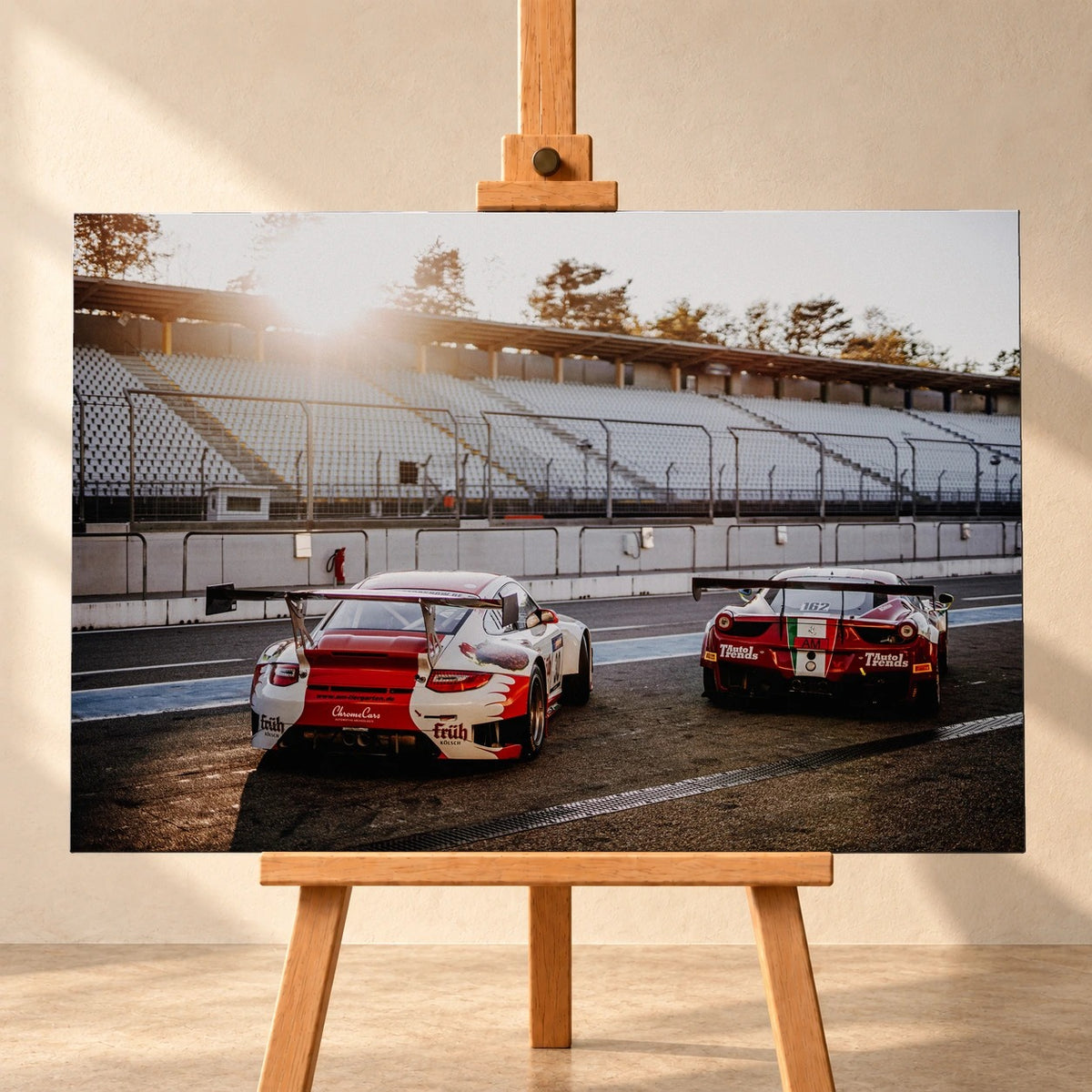 Frontale Visualisierung eines Leinwandbilds im Querformat auf einer Holzstaffelei vor heller Wand. Das Motiv zeigt einen rot-weißen Porsche 911 GT3 R links und einen roten Ferrari 458 GT3 rechts von hinten in der Boxengasse vor leeren, sonnenbeschienenen Tribünen.