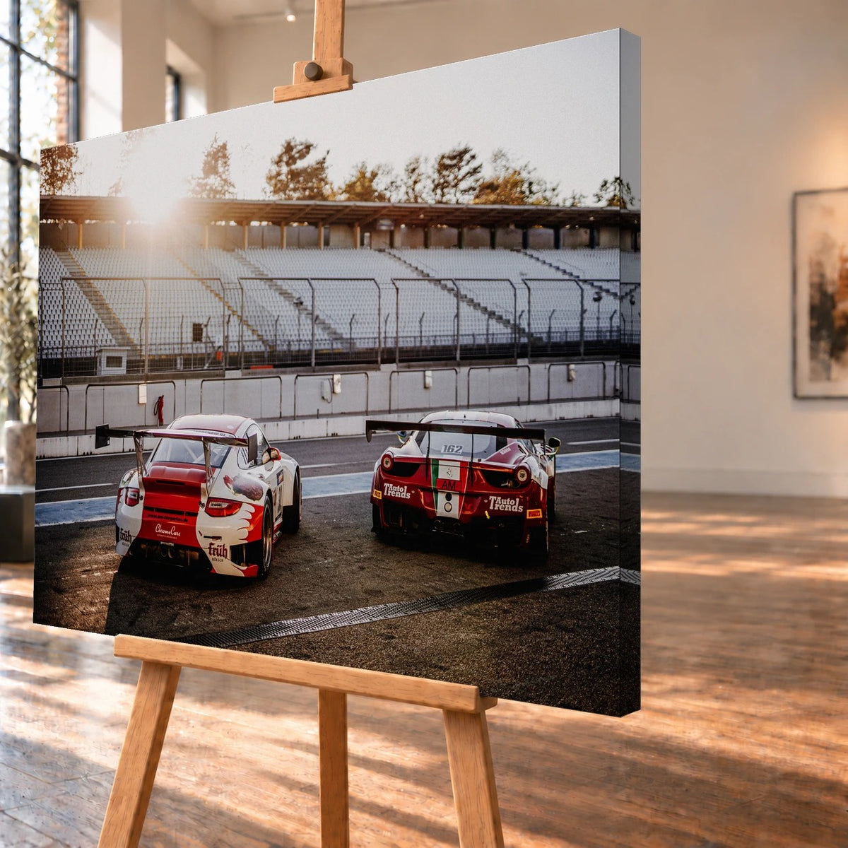 Schrägansicht eines Leinwandbilds als Visualisierung auf einer Holzstaffelei; das Motiv setzt sich über die sichtbare rechte Seitenkante fort. Porsche 911 GT3 R und Ferrari 458 GT3 stehen mit großen Heckflügeln in der Boxengasse, von hinten vor leeren Tribünen gezeigt.