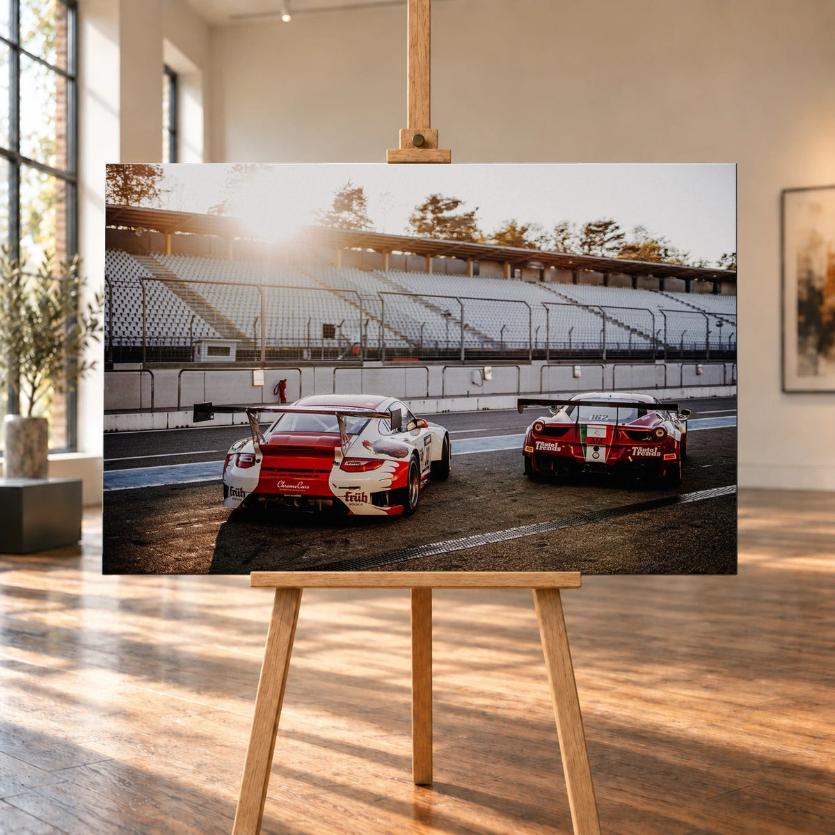 Raumvisualisierung mit einem querformatigen Leinwandbild auf einer Holzstaffelei in einem hellen Raum mit hohen Fenstern und Holzboden. Auf dem Bild sind Porsche 911 GT3 R links und Ferrari 458 GT3 rechts von hinten in einer sonnigen Boxengasse vor leeren Tribünen zu sehen.