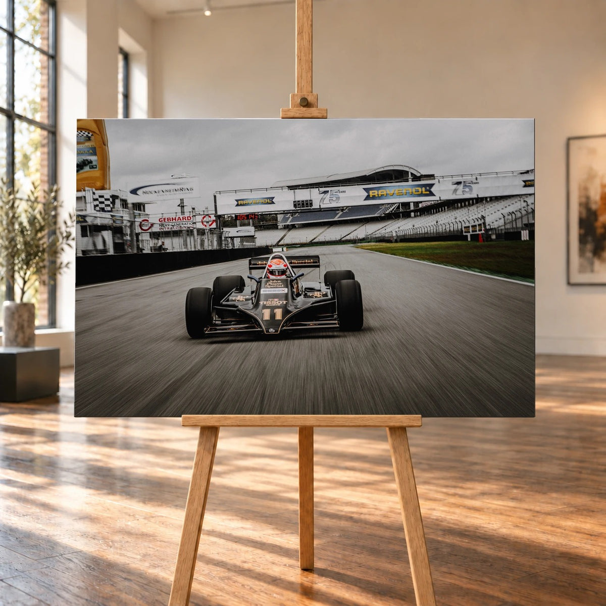 Raumvisualisierung mit einem querformatigen Leinwandbild auf einer Holzstaffelei in einem hellen Raum mit hohen Fenstern und Holzboden. Das Bild zeigt den schwarz-goldenen Lotus 87 mit Startnummer 11 auf der Rennstrecke vor den Tribünen.