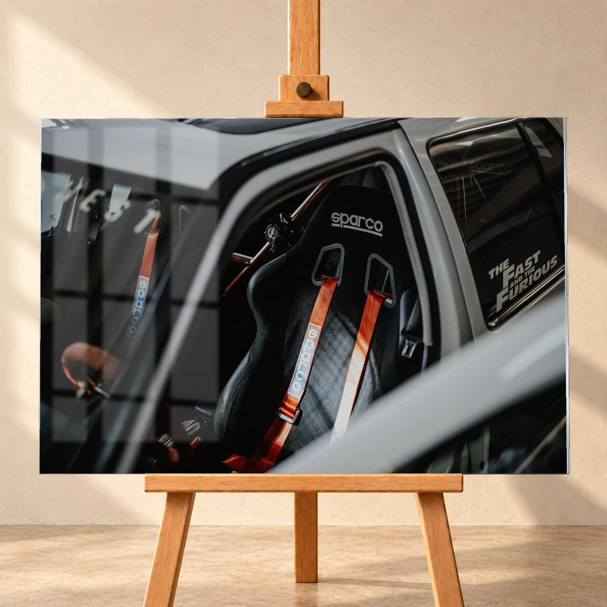 Frontal visualisiertes Acrylglas-Wandbild auf einer Holzstaffelei vor heller Wand, mit einer Fensterspiegelung auf der Bildfläche. Zu sehen ist der Innenraum von Jesses VW Jetta aus „The Fast and the Furious“, im Fokus ein schwarzer Sparco-Schalensitz mit roten Gurten.