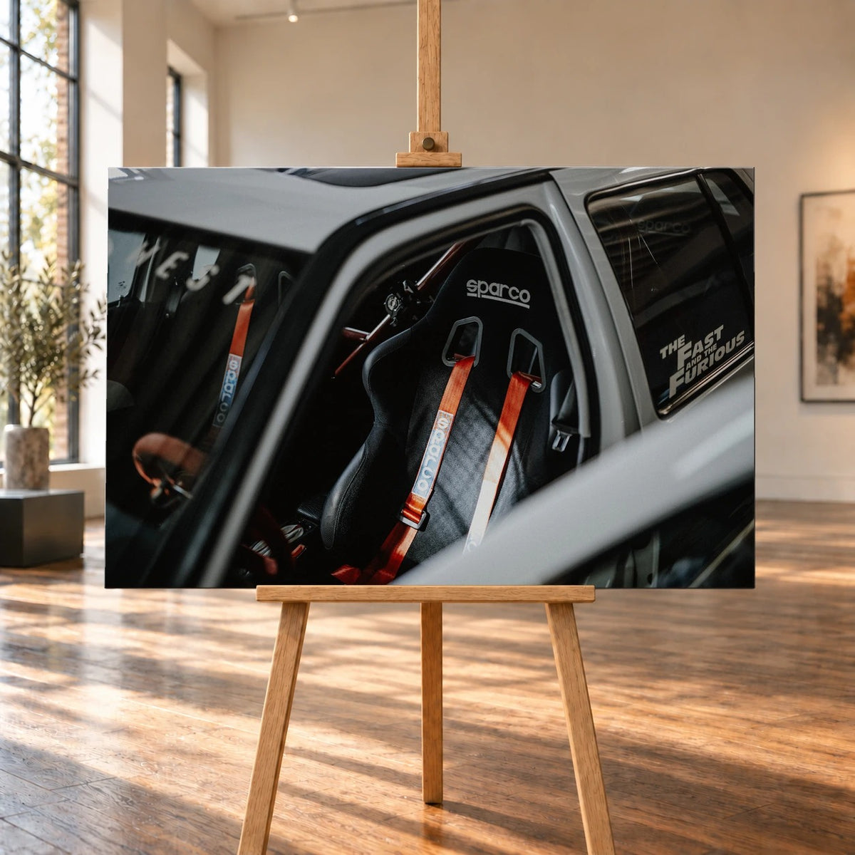 In einem hellen Raum mit großen Fenstern und Holzboden ist ein querformatiges Leinwandbild auf einer Staffelei visualisiert. Das Motiv blickt in Jesses VW Jetta aus „The Fast and the Furious“ und zeigt schwarze Schalensitze mit roten Gurten.