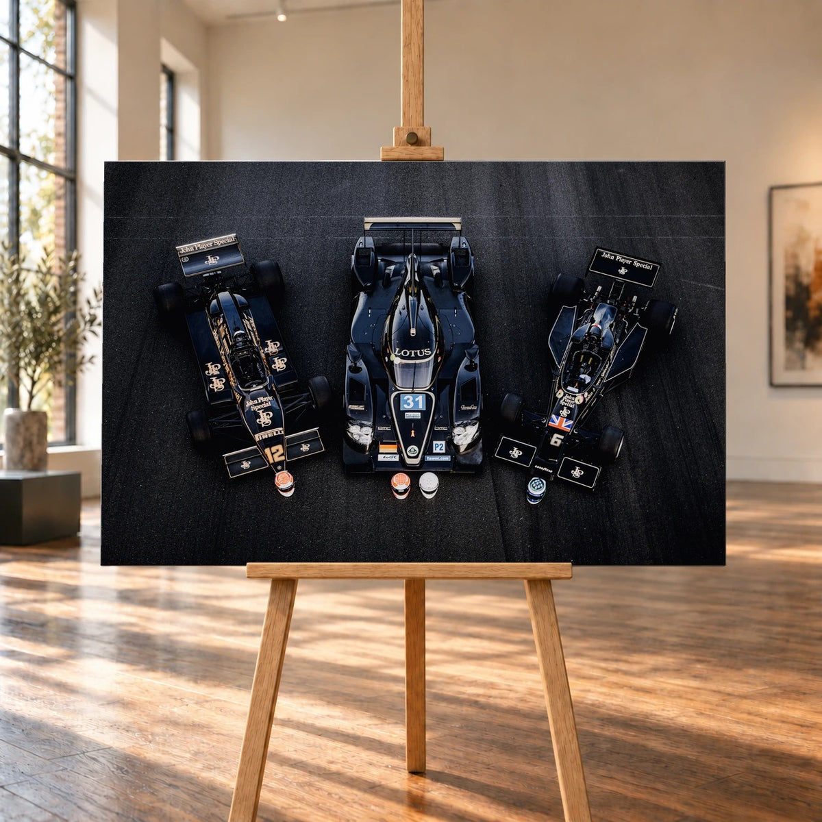 Raumvisualisierung eines querformatigen Leinwandbilds auf einer Holzstaffelei in einem hellen Raum mit hohen Fenstern und Holzboden. Auf dem Bild sind drei schwarz-goldene Lotus-Rennwagen in Draufsicht zu sehen, mit einem Sportprototyp zwischen zwei Formelwagen.