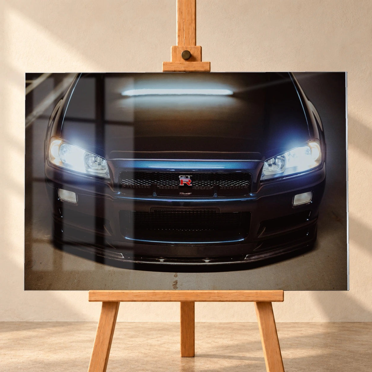 Frontal visualisiertes Acrylglas-Wandbild auf einer Holzstaffelei vor heller Wand. Es zeigt einen dunkelblauen Nissan Skyline GT-R R34 von vorn mit eingeschalteten Scheinwerfern und GT-R-Emblem. Eine Fensterspiegelung überlagert die linke Bildhälfte.