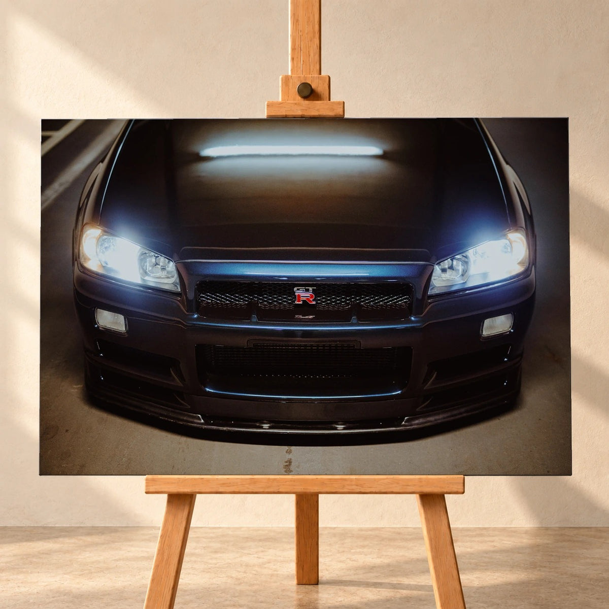 Visualisierung eines querformatigen Leinwandbildes auf einer Holzstaffelei vor heller Wand. Das Motiv zeigt die dunkelblaue Front eines Nissan Skyline GT-R R34 mit eingeschalteten Scheinwerfern und GT-R-Emblem im Kühlergrill.